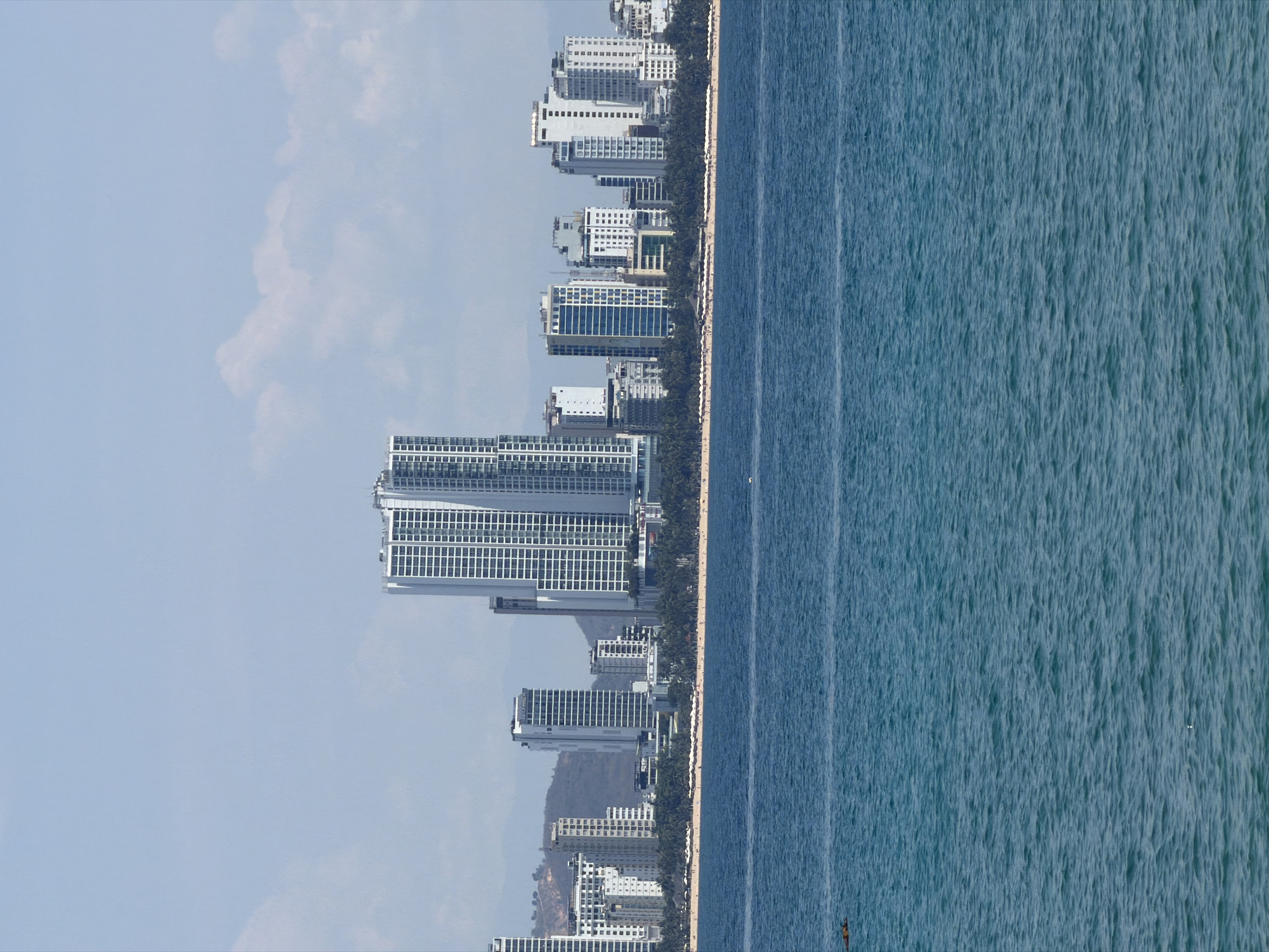 The image shows a coastal cityscape in Nha Trang Ward, Vietnam, viewed from a perpendicular orientation. Tall, modern buildings line the coast next to a beach with palm trees. Adjacent to the buildings and beach is an expanse of blue water. The other side of the image shows a light blue sky.