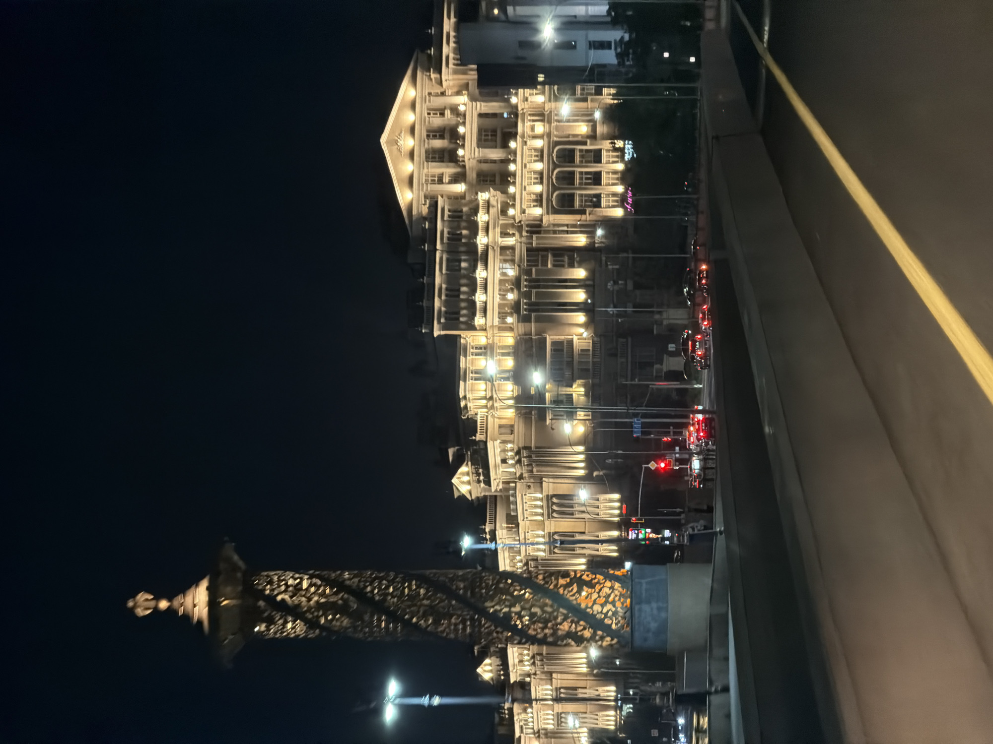 The image depicts a nighttime view of a city street in Astana, Kazakhstan, featuring a prominent, ornate column on the left and a large, illuminated building in the background. The column, made of a dark material with a spiraling, intricate pattern, is topped with a sculpted element. Streetlights illuminate the scene. The building is a multi-story structure with classical architectural details, adorned with numerous lit windows and decorative lighting that highlights its facade. Cars are visible on the street in front of the building, with their headlights and taillights illuminated. A traffic light shows red. The foreground shows a road or bridge with a yellow line marking. The sky is dark.