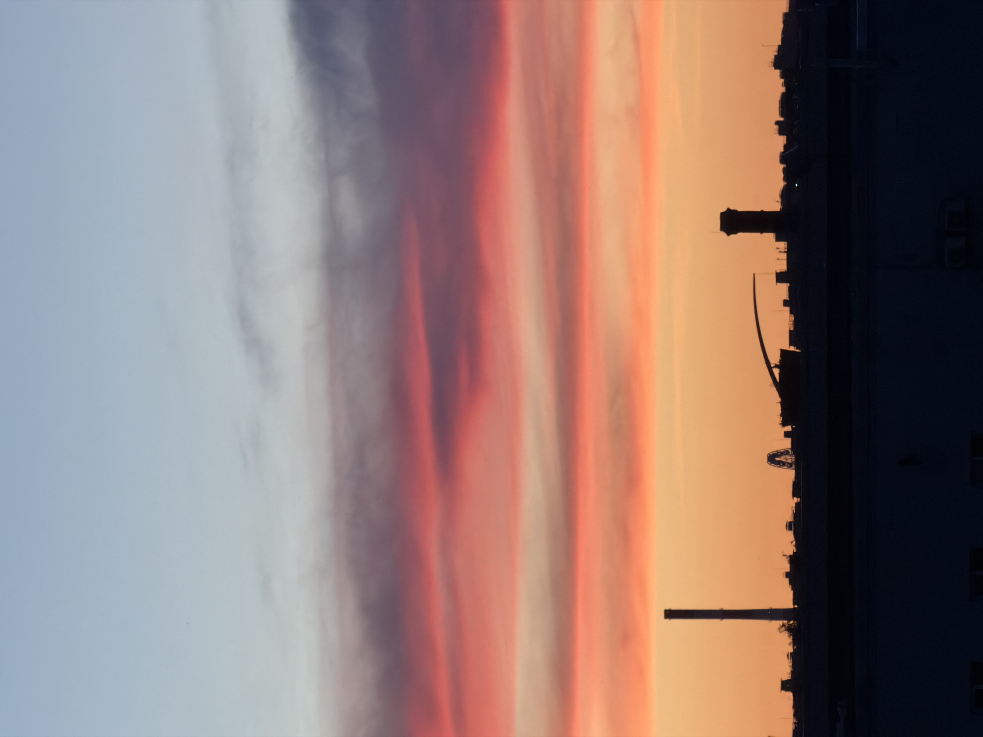 The image depicts a sunset over an industrial cityscape. The sky displays a gradient of colors, transitioning from pale blue at the top to vibrant oranges and pinks as it meets the horizon, with distinct bands of clouds colored by the setting sun. Silhouetted against this colorful sky is a cityscape featuring various structures, including tall chimneys, buildings, and what appears to be a Ferris wheel. In the foreground, a large, dark building dominates the lower portion of the frame, obscuring the base of the cityscape and suggesting a layered perspective of urban development. The overall impression is one of an industrial urban environment bathed in the dramatic light of dusk.