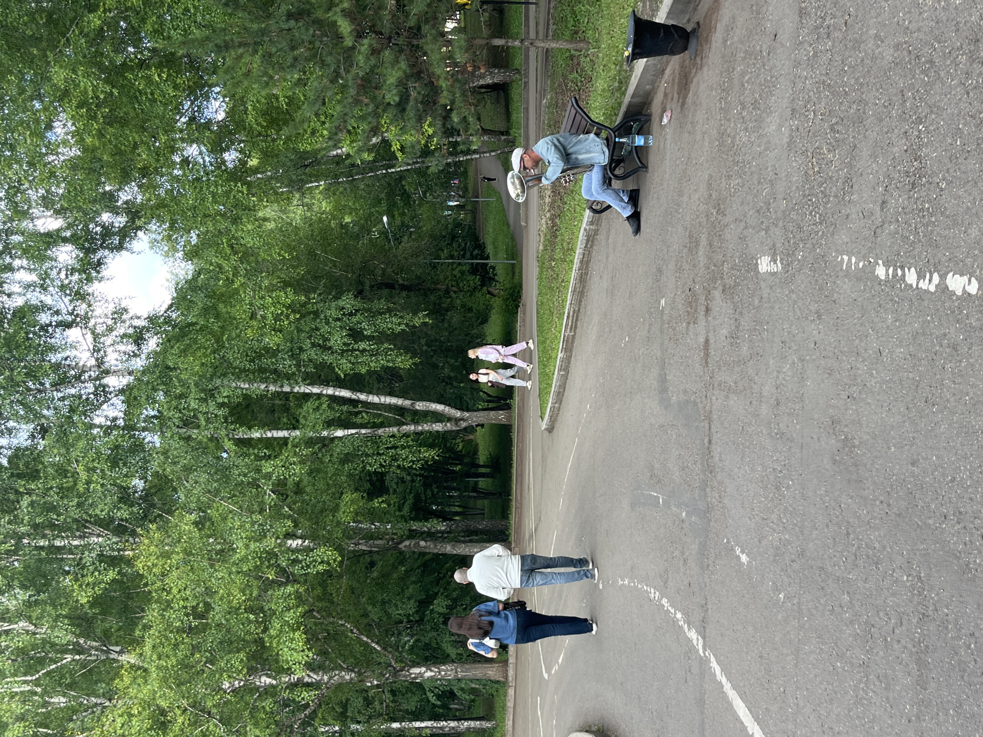 The image depicts a park scene with a paved path winding through a wooded area. In the foreground, two individuals, a woman in a blue denim shirt and dark pants and a man in a white shirt and jeans, walk away from the camera on the path. To the right, a man wearing a light blue denim jacket and jeans is seated on a dark bench, playing a large brass instrument with a bell. He is wearing a white cap. A bottle of water and a trash can are located near the bench. In the background, several other individuals are walking on a parallel path, and a lamp post is visible among the trees. The environment is characterized by lush green trees with white trunks, typical of birch trees, and a grassy area bordering the path. The overall impression is that of a peaceful, outdoor recreational space. The location is the city of Oktyabrsky, Russia.
