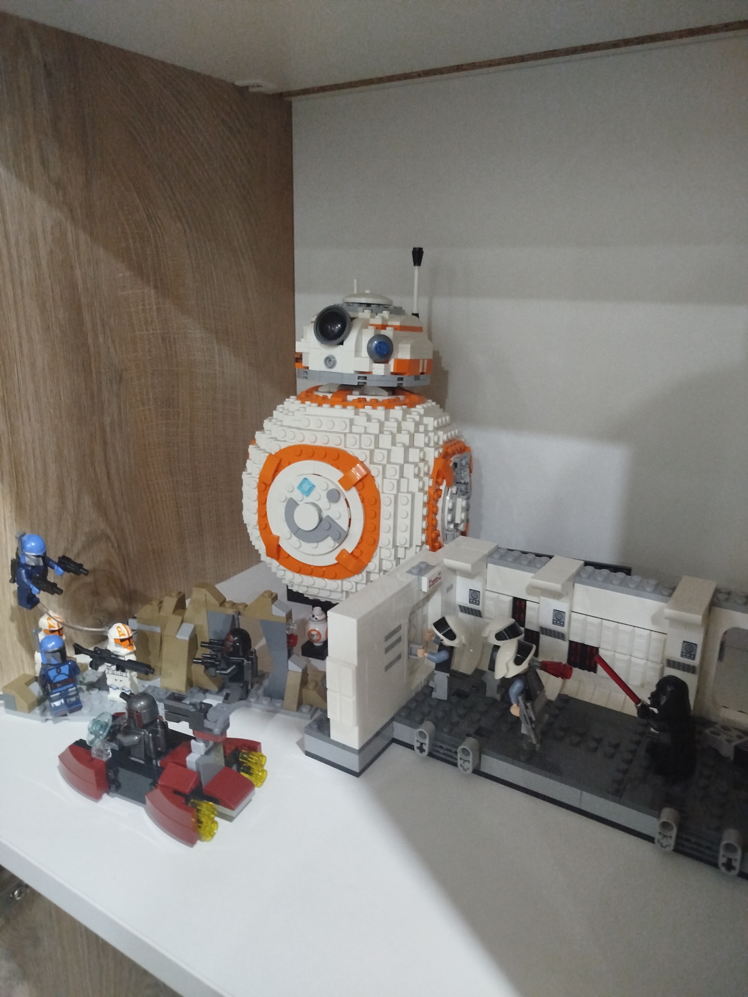A Star Wars Lego display is set up on a white shelf. On the left side of the display, there is a section of tan Lego bricks resembling terrain, with several Lego figures of Rebel soldiers, some in blue and orange uniforms, holding weapons. In front of them is a red and gray Lego vehicle with two figures, one in a Mandalorian-like suit. Dominating the center of the display is a large Lego model of the droid BB-8. To the right of BB-8 is a white Lego structure that appears to be a hallway or interior section, with several Lego figures of Imperial stormtroopers and a Darth Vader figure holding a red lightsaber. The background shows a wooden textured wall on the left and a plain white wall on the right. There are no indications of the city of Mirny, Russia, in this image.