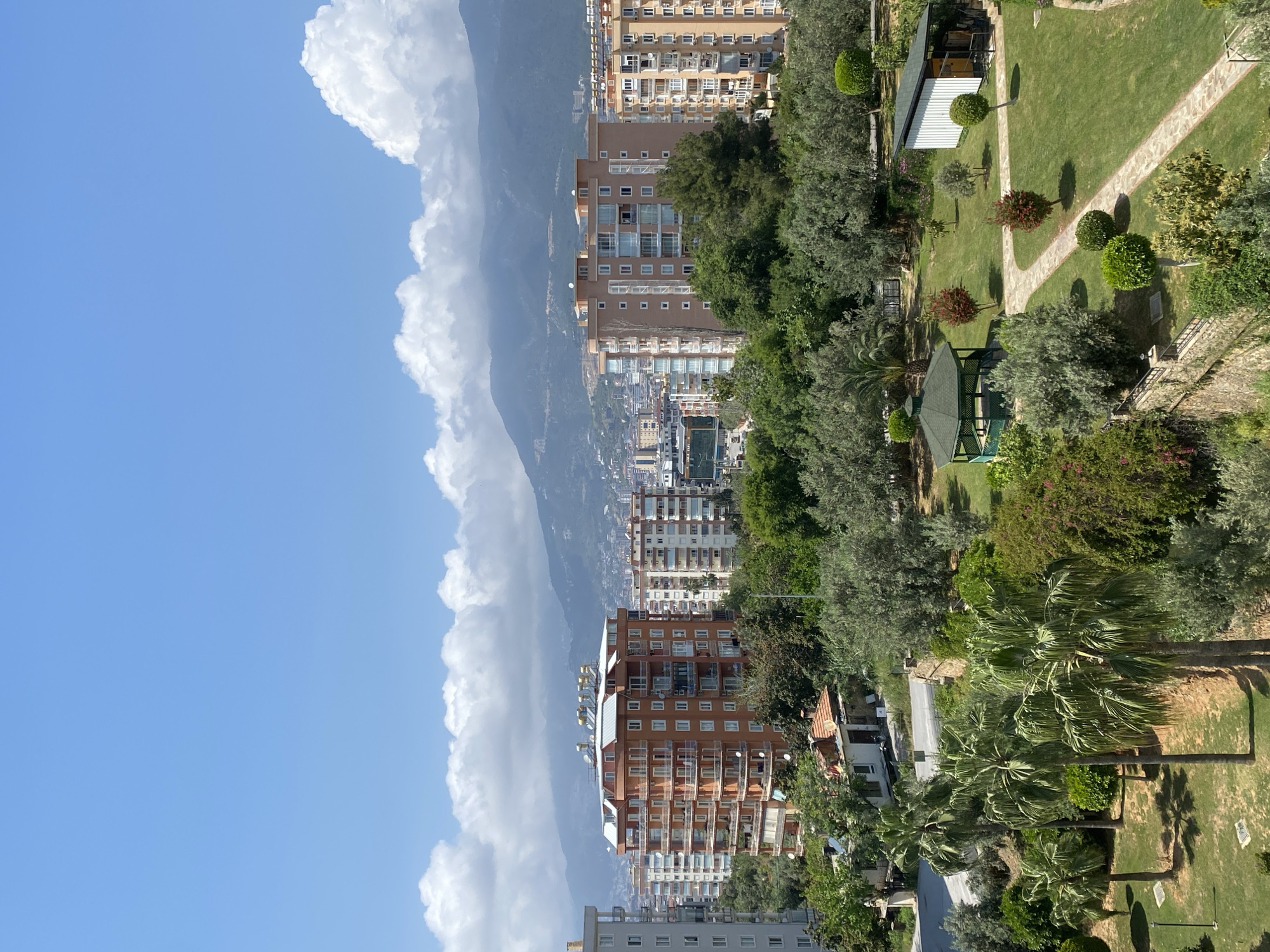 This panoramic image, taken in Alanya, Turkey, presents a layered view encompassing a vibrant park, a bustling city, and a majestic mountain range under a clear blue sky.
In the foreground, a lush green park unfolds with manicured lawns, scattered trees of various types, including prominent palm trees, and two meandering stone-paved pathways. A green-roofed wooden gazebo is nestled among the foliage, alongside a smaller building with a white roof. Distinct shadows indicate bright sunshine.
The midground showcases a dense urban environment featuring numerous multi-story residential buildings. These structures, primarily light brown and white, display many windows and balconies, with some buildings equipped with satellite dishes. Glimpses of city streets and additional infrastructure can be seen between and beyond these buildings.
Dominating the background are large, greyish-green mountains, partially obscured by a dramatic formation of white cumulus clouds that line their peaks. The sky above the mountains is a brilliant, unblemished blue.
The scene depicts a clear and sunny day, likely in the mid-morning to late afternoon, given the strong illumination and visible shadows. There are no people or specific activities visible. The image conveys a serene yet vibrant atmosphere where urban life harmoniously meets natural landscapes. No visible text is present.
