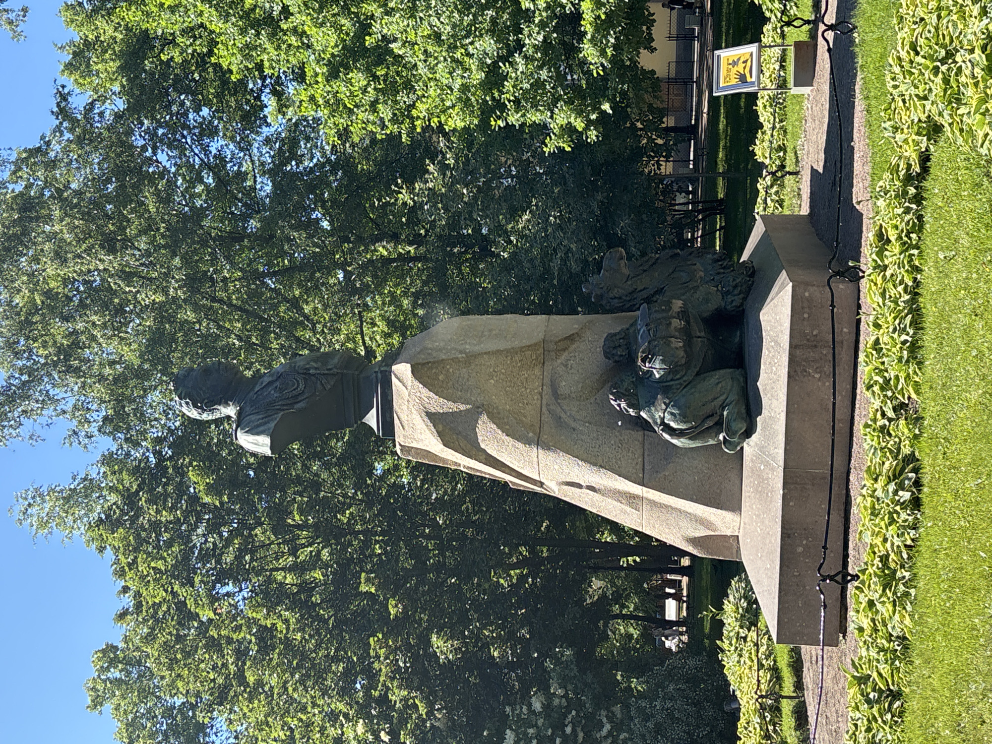 This is a full shot of a monument in a park setting. The monument is made of stone and bronze, featuring a bust of a person on top of a pedestal. The bust is facing left and appears to be a man with a formal hairstyle. Below the bust, the pedestal is adorned with bronze figures. The scene is outdoors, with lush green trees and grass surrounding the monument. The lighting suggests it is daytime, likely sunny, as evidenced by the bright sky and the shadows cast by the monument and trees. The overall atmosphere appears peaceful and serene, typical of a public park.
There is a yellow sign with black symbols to the right of the monument, attached to a pole. The sign has an image of a person's hands and a warning symbol. Further back, a dark metal fence encloses a pathway and a building with a light-colored facade. The foliage in the foreground and background is vibrant, indicating a pleasant season, likely spring or summer. The monument itself is weathered, suggesting it has been a part of the park for some time. The image is taken from a slightly low angle, emphasizing the monument's presence.