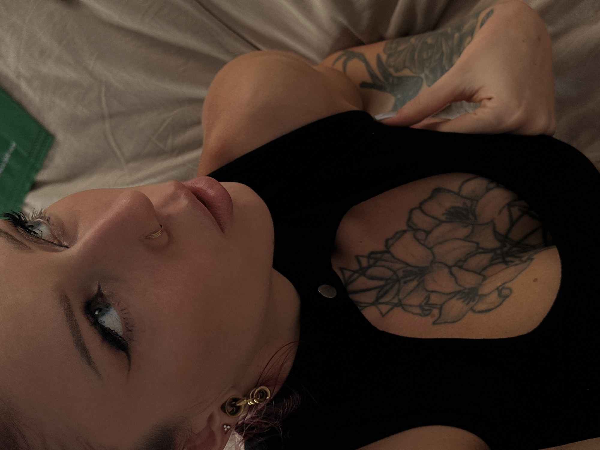 A person is lying on a bed with a gray sheet. The person has a black top with a cutout in the chest area revealing a flower tattoo. The person has tattoos on their upper arm. The person has a nose ring and a large earring in their left ear. A green object is visible on the left edge of the frame. The location is not specified.