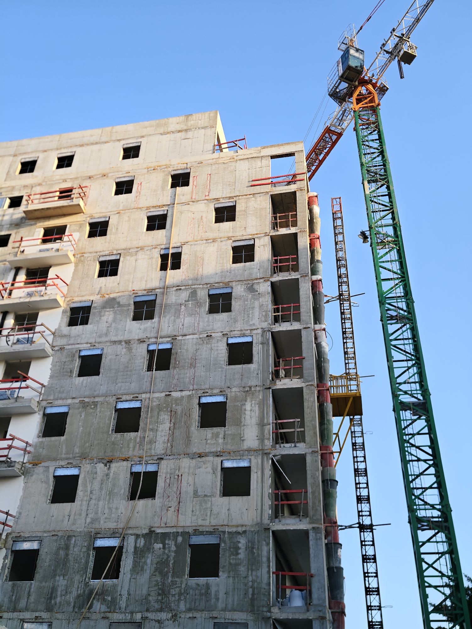 An unfinished concrete apartment building under construction is shown in Petah Tikva, Israel, against a clear blue sky. A large green tower crane is positioned to the right of the building, with its boom extending over the top. The building has multiple floors, each with several window openings that appear empty or boarded up. Red safety railings are visible on some of the balconies and around unfinished sections of the building. A long rope or cable hangs vertically down the side of the building. The scene depicts an active construction site with ongoing building work.