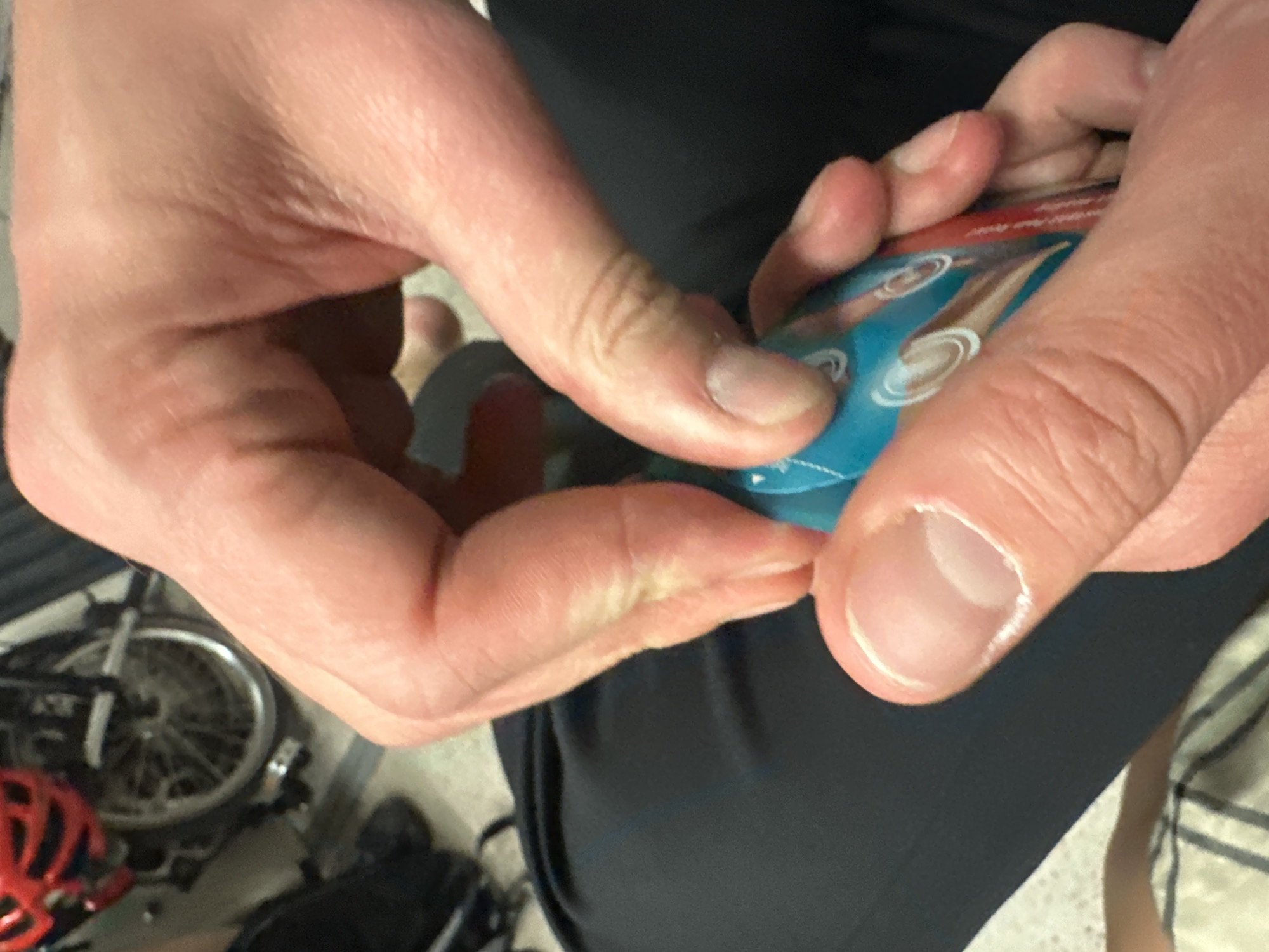 A close-up shot shows two hands opening a small, blue and red packet. The person is wearing dark clothing. A bicycle and red helmet are partially visible in the lower left corner of the frame. The location is not discernible.