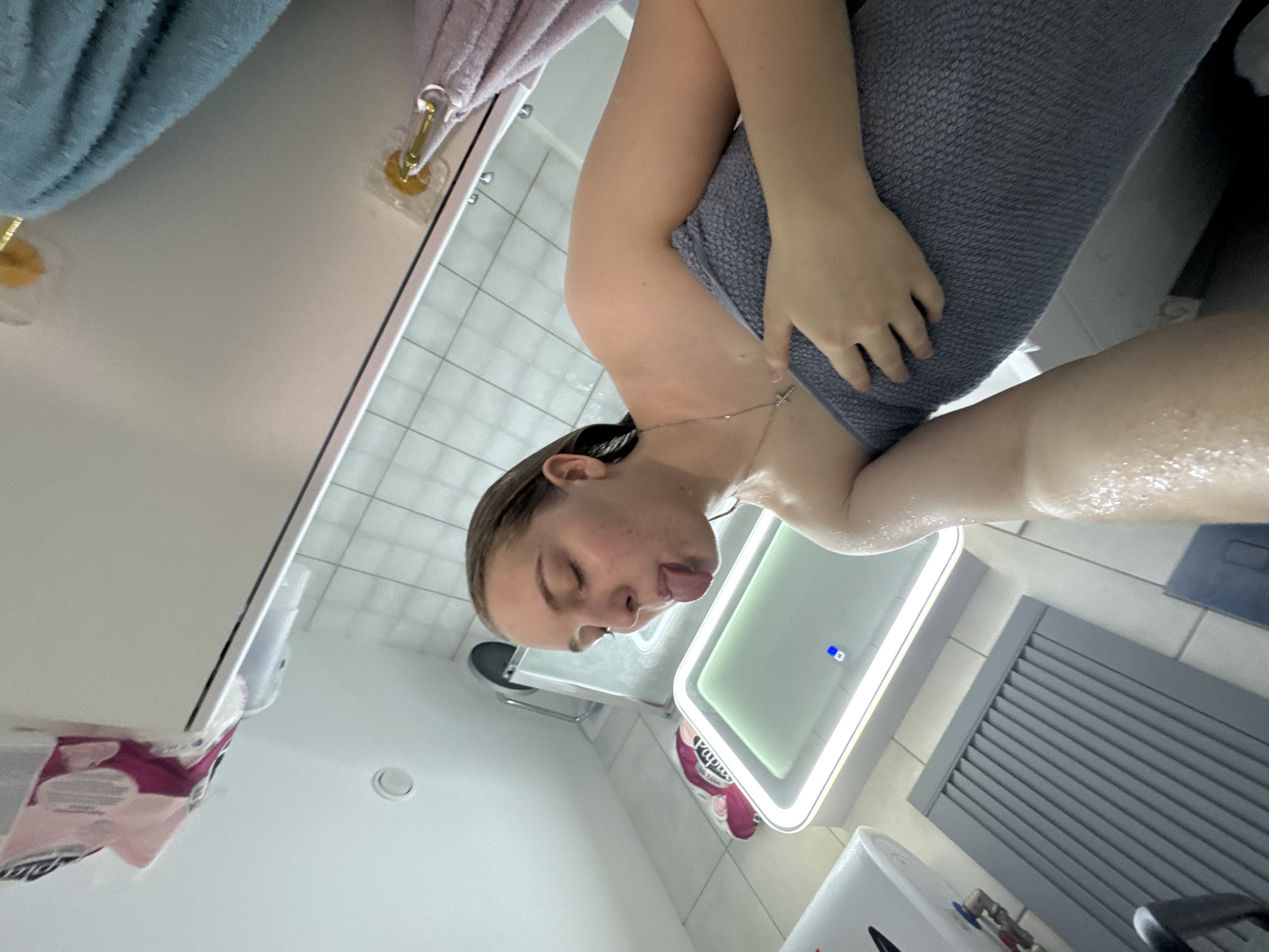 The image depicts a young adult female in a bathroom setting, likely after showering. She has wet, light brown hair pulled back from her face, and water droplets are visible on her skin. She is wrapped in a grey towel that covers her torso. Her eyes are closed or heavily squinted, and she is extending her tongue from her mouth. She wears a silver chain with a small cross pendant. The perspective is a low-angle selfie, with a wet arm partially visible in the foreground.
The background shows white tiled walls with light grout and a white ceiling. A rectangular mirror with an illuminated frame and a small blue indicator light is central in the background. To the left, a white wall-mounted water heater with visible red and blue pipe connections is present. A grey slatted floor drain cover is on the floor. In the upper right, multiple clear plastic suction cup hooks with gold-colored hangers are attached to a wall or shower partition, holding a blue towel and a pink/lavender towel. A roll of toilet paper in pink packaging is visible in a white holder near the floor on the left. A glass shower enclosure and what appears to be a fan or vent are partially visible behind the subject.
The specific location within the city of Perm, Russia, cannot be determined from the visual information provided in the image.