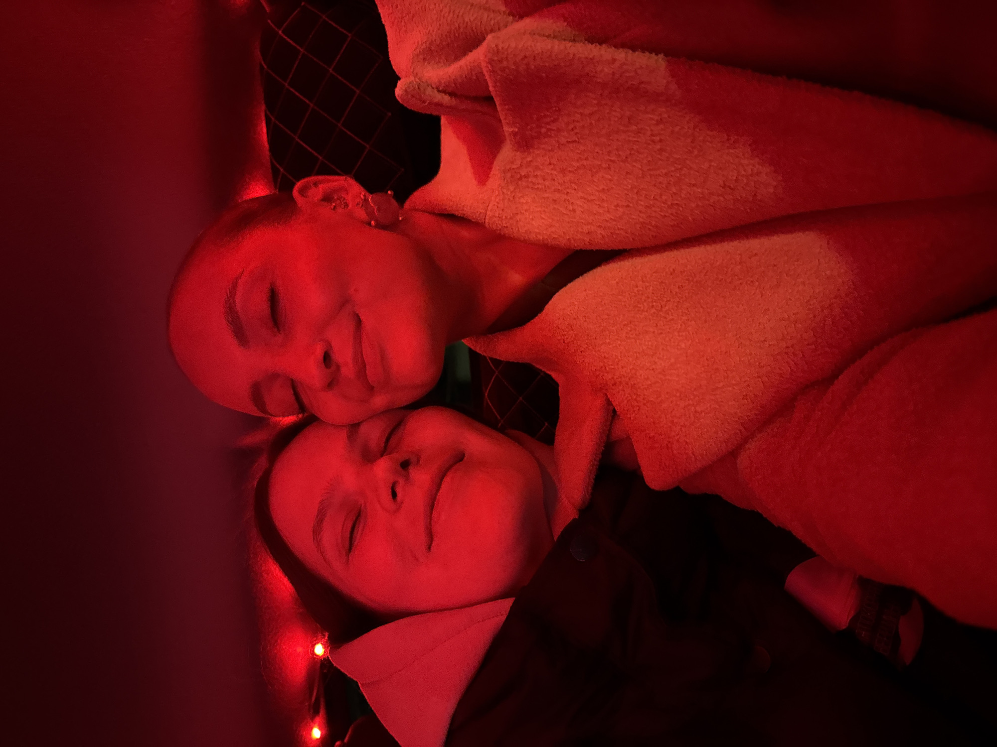 This close-up, top-down image captures two individuals, likely women or young adults, lying intimately together in an indoor setting in Chaikovsky, Russia. The scene is bathed in a predominant red light, creating a warm, cozy, and soft ambiance, possibly from string lights or special lighting.
The subjects are positioned with their heads close to each other, one slightly above and to the left of the other. Both have their eyes closed and exhibit soft, serene smiles, conveying feelings of peace, contentment, and affection. The upper individual is partially covered by a light-colored, soft, and possibly fuzzy blanket or garment, which wraps around their neck and chest. The lower individual wears a darker garment, possibly a jacket or shirt, over a lighter collar. Around the lower person's head and hair, small, glowing red string lights are visible, contributing to the festive or intimate atmosphere.
Behind the upper individual's head, a dark, textured surface with a grid or diamond pattern can be seen. The red lighting saturates the scene, casting deep shadows and highlighting the contours of their faces and the soft textures of their clothing. The time of day is not explicitly clear, but the artificial red lighting suggests it is evening or night. No specific weather conditions are discernible, as the setting is indoors. There is no visible text in the image.