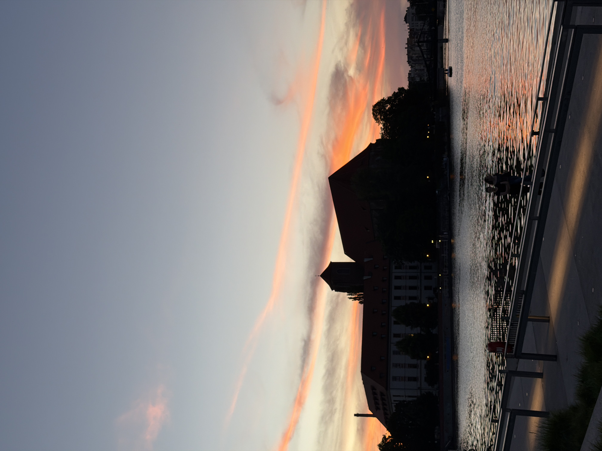 At dusk, the sky over an unknown city is painted with hues of orange and pink, with wispy clouds streaking across the pale blue expanse. A wide body of water, reflecting the vibrant sky, dominates the lower half of the frame. In the mid-ground, a silhouetted building with a red-tiled roof and a tall tower stands prominently on the opposite bank of the water. Trees partially obscure the building. A walkway with metal railings runs along the foreground, leading the eye towards the water. Two women are standing on the walkway, facing the water, their silhouettes dark against the illuminated sky. A small boat is visible on the water in the distance, and a bridge spans the waterway further down. The scene is peaceful and serene, capturing a moment of quiet beauty as day transitions into night.