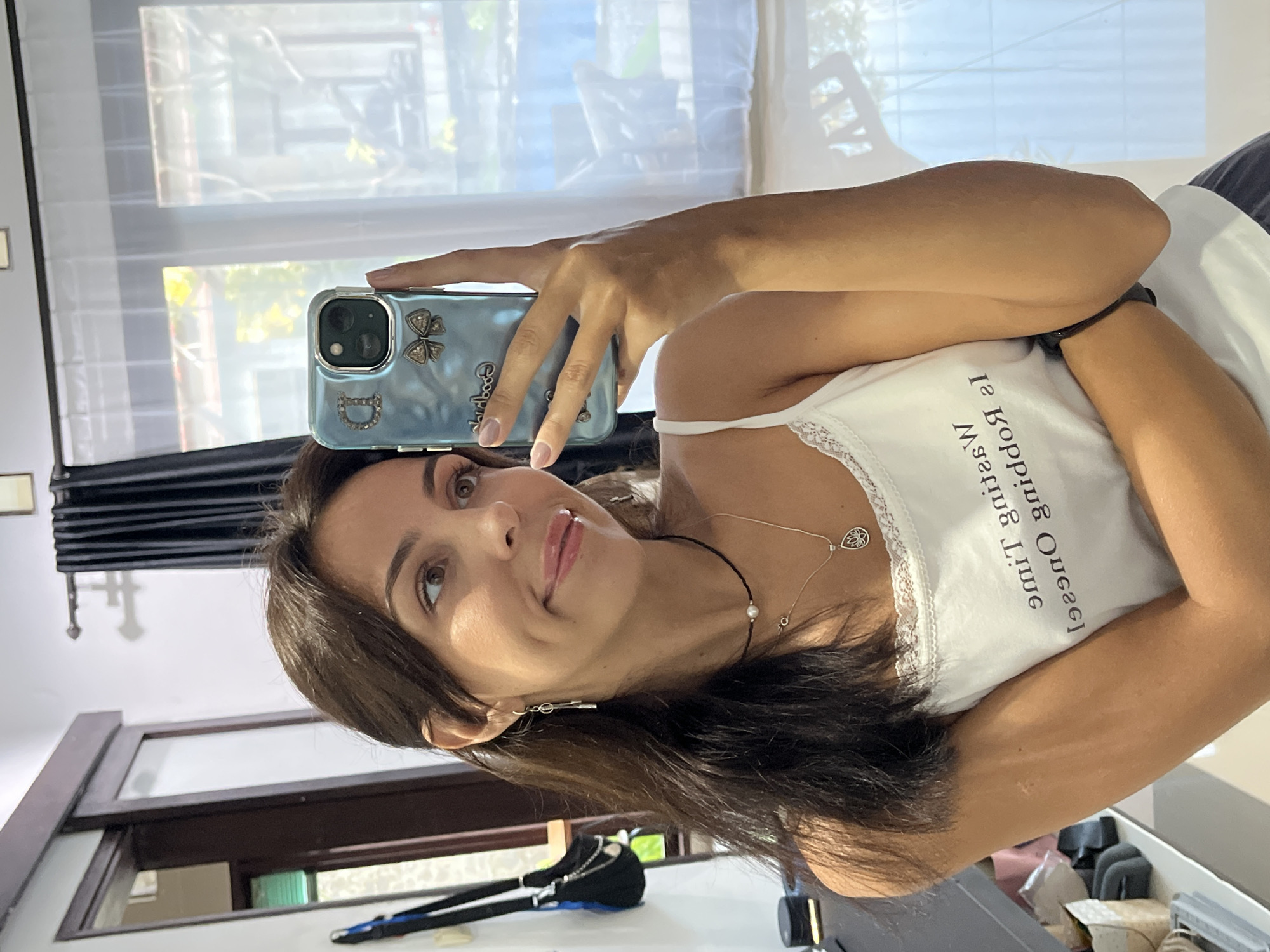 A woman is taking a selfie with a blue phone case. The case has decorations on it. She is wearing a white tank top with text written on it. She has brown hair and light skin. She's wearing two necklaces. The location appears to be inside a residence in Unknown, Indonesia, as evidenced by the interior design and the view from the window, which includes local foliage. The room has a window with white curtains and a black curtain. There are household objects and furniture visible in the background.