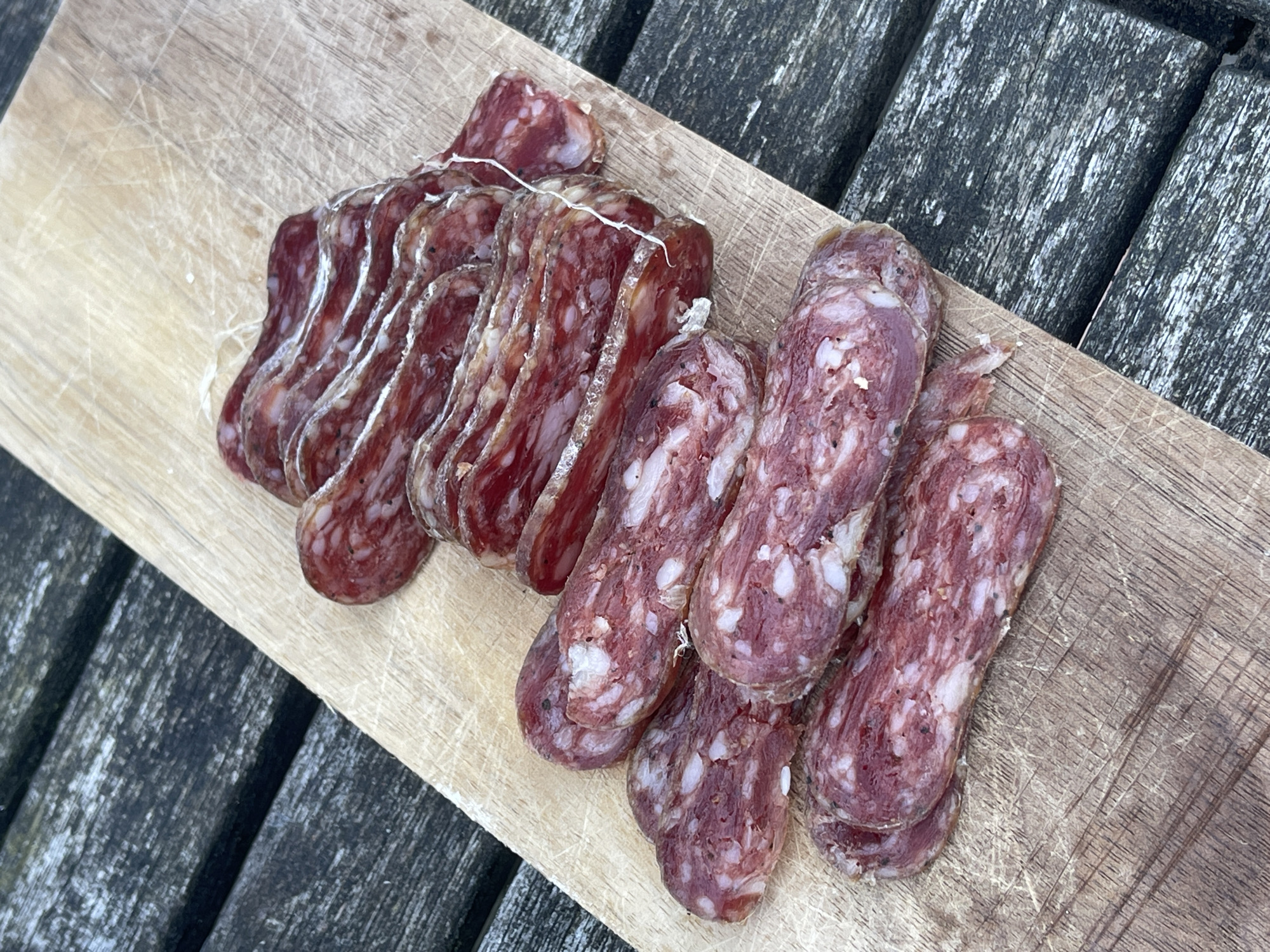 The image depicts a wooden cutting board on a weathered wooden surface, possibly an outdoor table. The board holds two portions of salami; one is sliced and fanned out, revealing thin, marbled pieces. The other is composed of four uncut, small salamis, slightly overlapping. The image does not contain sufficient information to place it in Maidstone, United Kingdom, or to identify any individuals or interactions. The focus is solely on the arrangement of the food item.