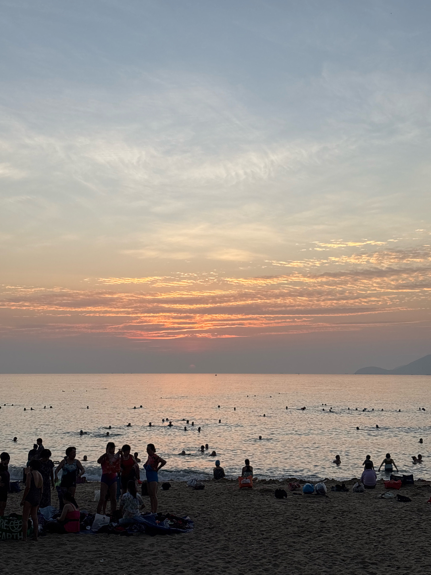 A group of people are enjoying a sunset at a beach in Nha Trang, Vietnam. The sky is filled with soft hues of orange, pink, and blue, with wispy clouds scattered across the horizon. The sun is low in the sky, casting a warm glow over the ocean. Numerous people are in the water, swimming and playing, while others are gathered on the sandy shore, relaxing and socializing. Some individuals are wading in the shallow water, while others are further out. Beach towels and bags are spread out on the sand. The overall atmosphere is one of peaceful enjoyment of a beautiful natural setting.