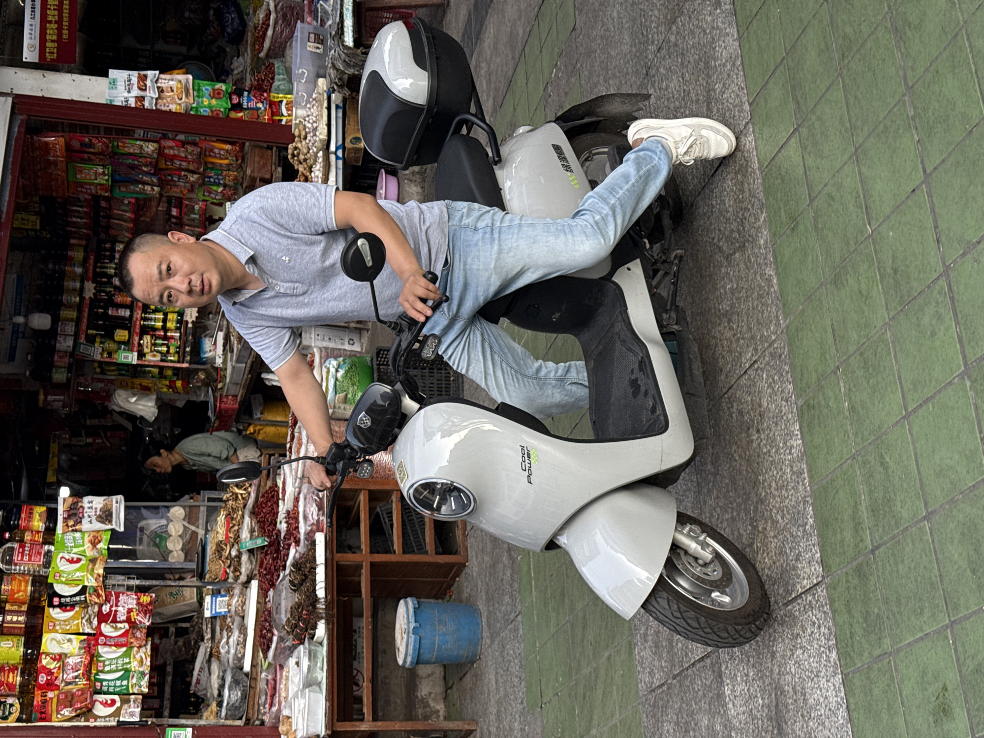 An upright, eye-level shot captures a man posing on a white electric scooter in what appears to be a busy street in Jiangjin, China. The man, with short dark hair, is wearing a light gray polo shirt, light blue jeans, and white sneakers. He is leaning back on the scooter's seat with his right leg casually extended over the back, while his left hand rests on the handlebar. His expression is neutral, looking directly at the camera.
The scooter is white with black accents and a black seat. A gray and black helmet is stored on a rack at the rear of the scooter. On the side of the scooter, the words "Cool Power" are visible in green, written vertically.
In the background, to the man's left, is the entrance of a shop. The shop is filled with various packaged goods, including snacks, drinks, and what appears to be dried food items like chilies and roots hanging from displays. Shelves inside the shop are packed with colorful merchandise, and a person is faintly visible deeper inside. Outside the shop, there are also wooden display shelves and a blue barrel.
The ground consists of a green-tiled sidewalk section to the right, and a darker, textured surface, possibly asphalt or concrete, to the left. The scene suggests a daytime setting, possibly overcast, given the soft lighting and lack of harsh shadows. Visible Chinese characters appear on signs and product packaging within the shop, consistent with the location. A small QR code is also visible on a shelf inside the shop.