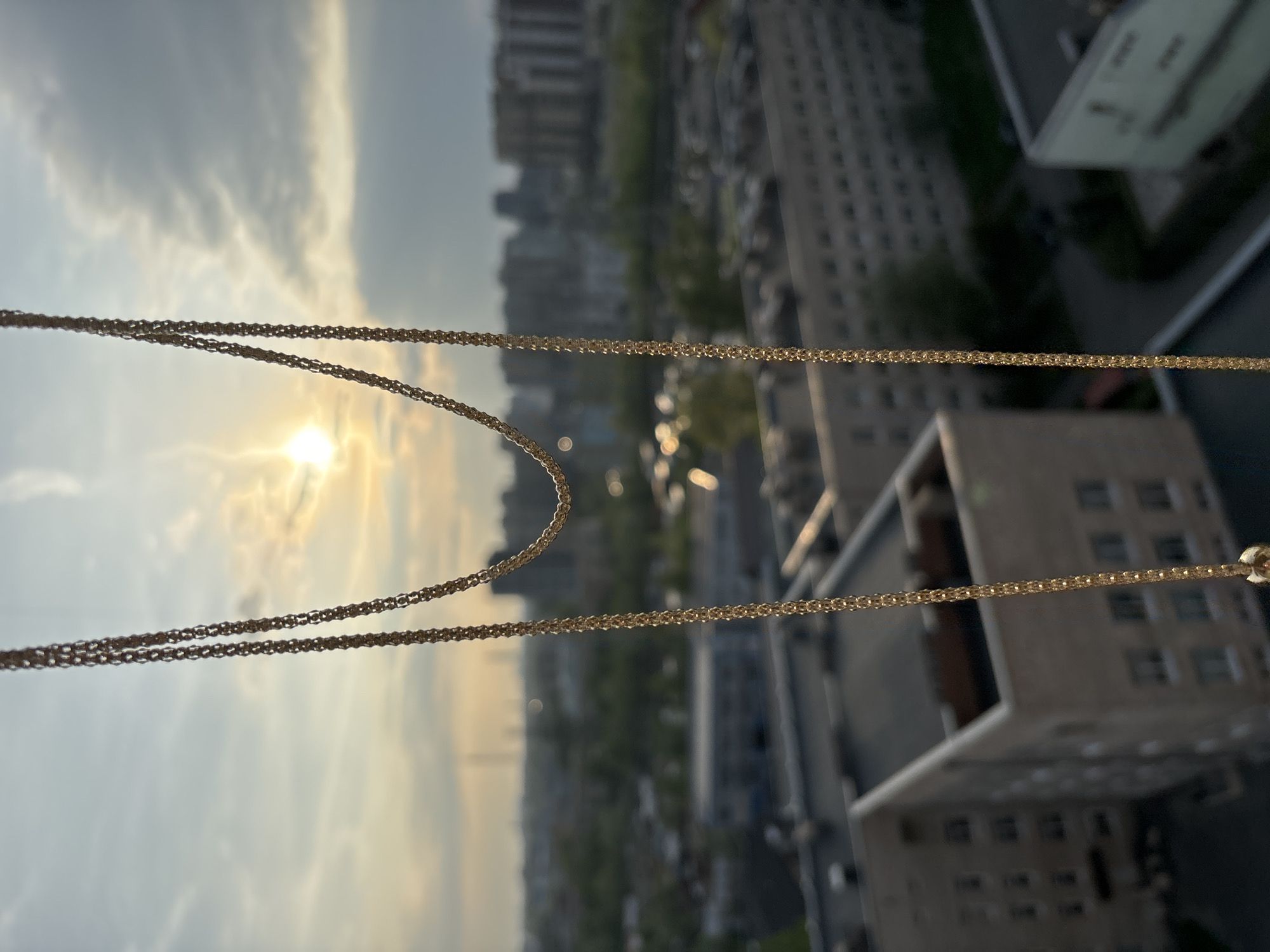 A medium-angle, blurred, rotated indoor shot shows a thin gold chain against a cityscape. The chain is in focus, contrasting with the blurred city buildings in the background. The sun is visible, shining through the upper portion of the chain. No people are visible. The location is a city, with numerous buildings, but the city is not identifiable.