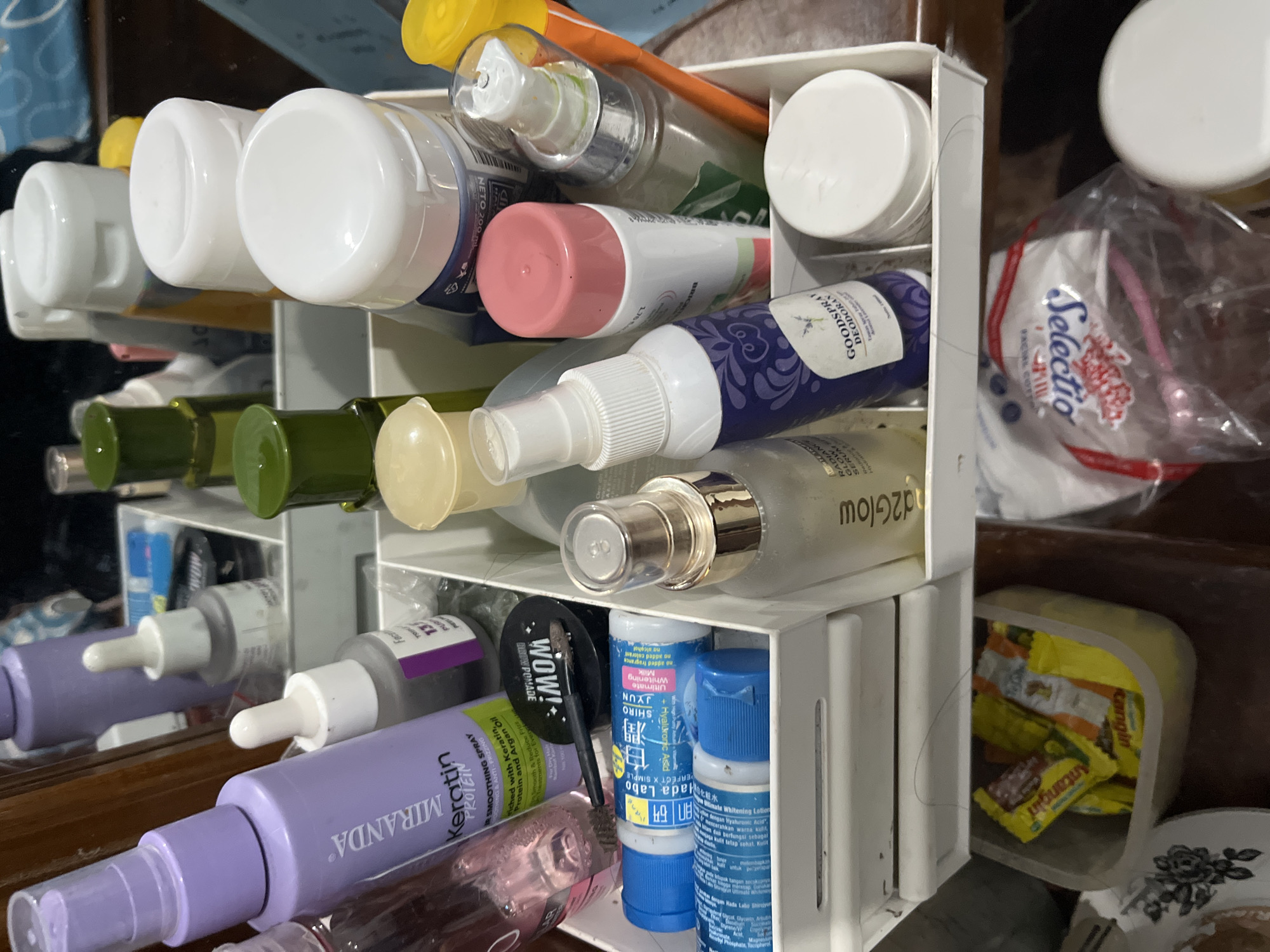 The image shows a close-up view of a white organizer filled with various beauty and skincare products. The organizer is placed on a wooden surface, possibly a vanity or desk. In the background, a mirror reflects some of the items and the surroundings.
Prominently featured in the organizer are several bottles and containers. On the left, a tall purple bottle labeled "MIRANDA Keratin Protein" is visible, along with a smaller glass bottle with a dropper. Further back, a green-capped bottle and several white-capped containers are arranged. A blue and white tube labeled "HADA LABO" is also present.
In the foreground and center, a spray bottle with a purple and white label that reads "GOODSPRAY Deodorant" is visible. Next to it is a pink-capped aerosol can. To the right, there are more white-capped containers and a yellow-capped bottle. A clear glass bottle with a spray nozzle is also in the mix.
Beneath the organizer, a clear plastic drawer contains several individually wrapped yellow packets, possibly snacks or tea. To the right of the organizer, a small portion of a white container and a red and white packaged product, possibly cotton pads, are visible.
The overall scene suggests a personal care and beauty collection. The lighting appears to be artificial, and the focus is on the contents of the organizer. There is no indication of the city of Glagah Kulon, Indonesia, in the image itself.