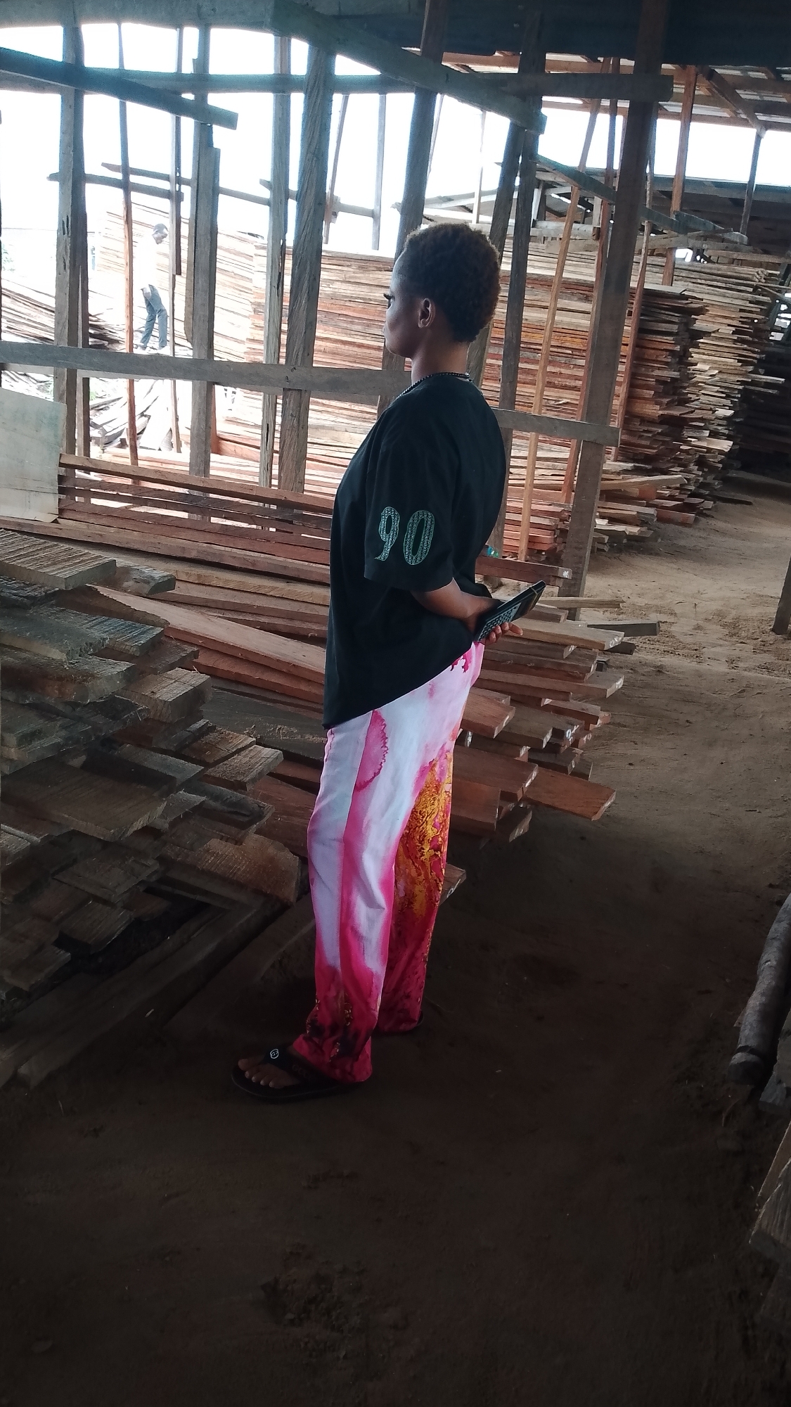 The image shows a woman standing in what appears to be a lumberyard in Asubi, Nigeria. The woman is wearing a black t-shirt with the number "90" on it, along with white and pink pants, and black sandals. She is looking to the right and holding a small black object in her hands. Stacks of lumber are visible around her. In the background, structural beams of the building are visible, and a person can be seen further in the distance behind the beams. The ground appears to be dirt.