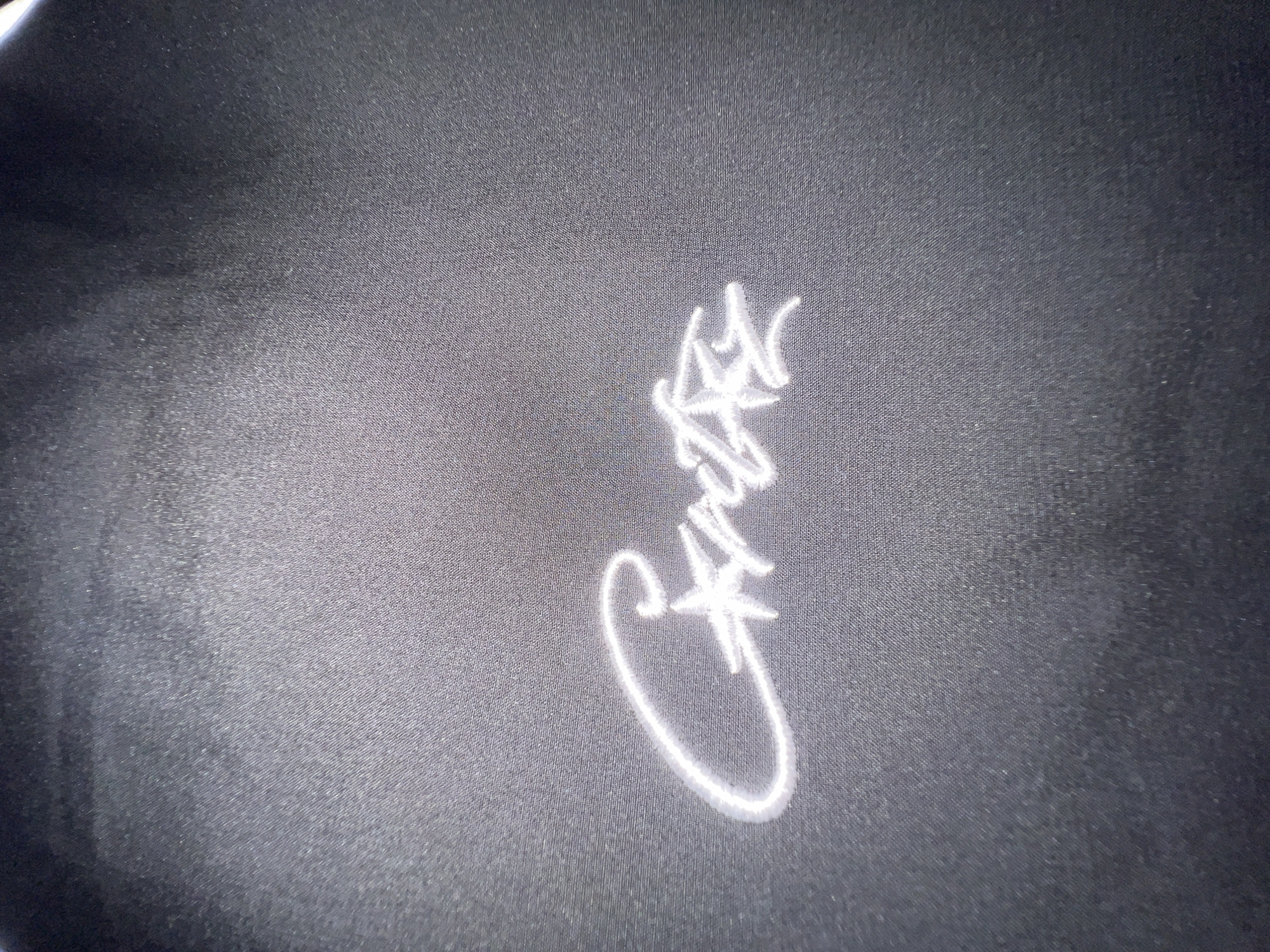 The image shows a close-up of white embroidered text on a dark, possibly black, fabric surface. The text is a cursive script, appearing to spell "Cailla". The background fabric has a visible weave pattern. The location in Krasnoyarsk, Russia is not apparent from the image. There are no people visible.