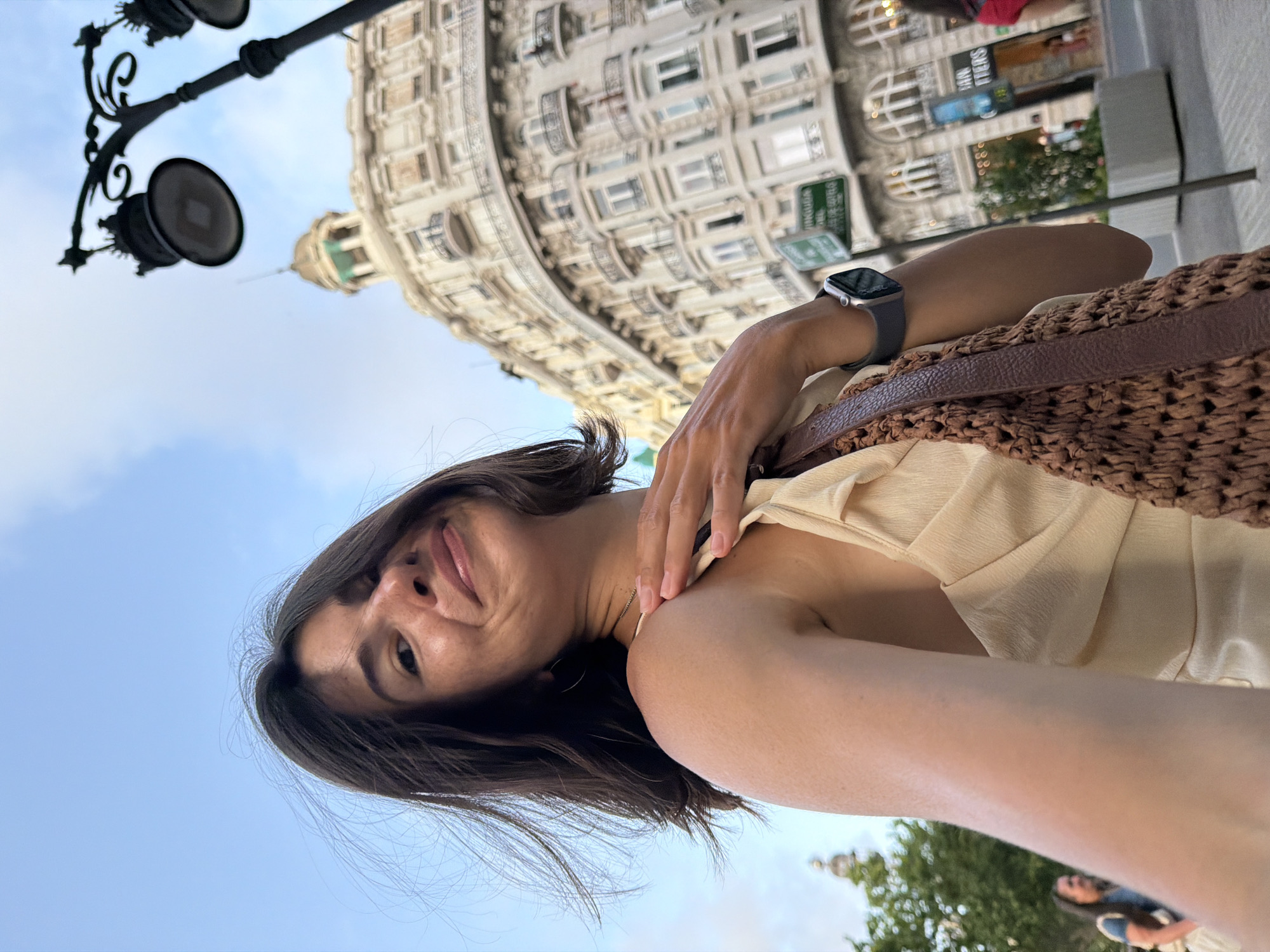 A medium shot captures a woman with dark hair in a selfie, looking towards the camera with a smile. She is wearing a beige dress and a brown crocheted bag slung over her shoulder. Her left hand is placed on her right shoulder. In the background, a grand, ornate building with balconies and numerous windows dominates the scene. A lamppost with two lights is visible in the upper right corner, and a blue sky with scattered clouds forms the backdrop. The setting appears to be a city street with some greenery and other people in the distance, suggesting an outdoor urban environment.