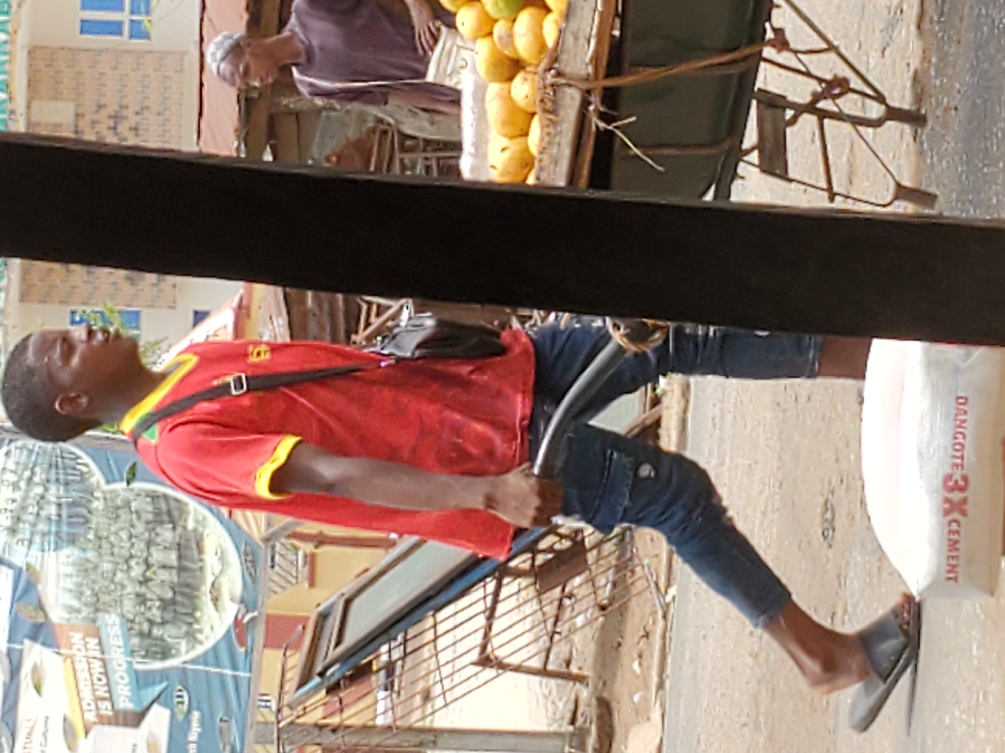 The image depicts a scene in Mararaba, Nigeria. A young man wearing a red t-shirt and jeans is pulling a cart loaded with mangoes. An older man wearing a purple robe is seen behind the cart. The cart is tied with rope. Next to the young man is a bag of Dangote 3X Cement. The setting appears to be an urban street with buildings and a billboard visible in the background.