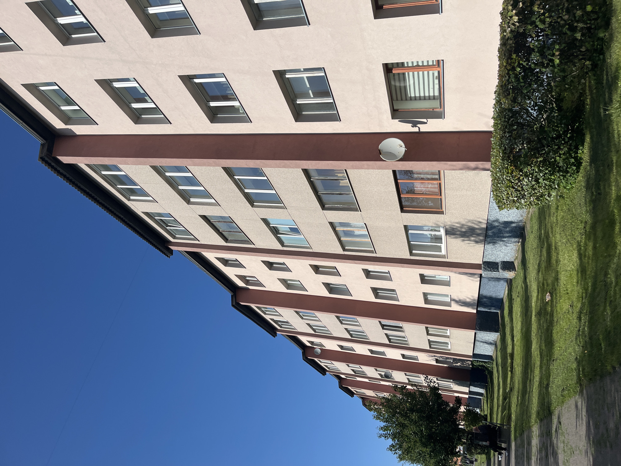 The image shows a multi-story residential building with a light pink facade and darker pink vertical accents, located in Kohtla-Järve linn, Estonia. The building has multiple rows of windows, most of which appear to be white-framed. A dark, corrugated roofline is visible at the top of the building. The building is situated next to a well-maintained green lawn with several trees and hedges. A paved walkway runs along the left side of the lawn, and a dark bench with two figures is present on the walkway. The sky is clear and bright blue. There are satellite dishes attached to the facade of the building. The lower portion of the building's facade has a textured, dark blue base.