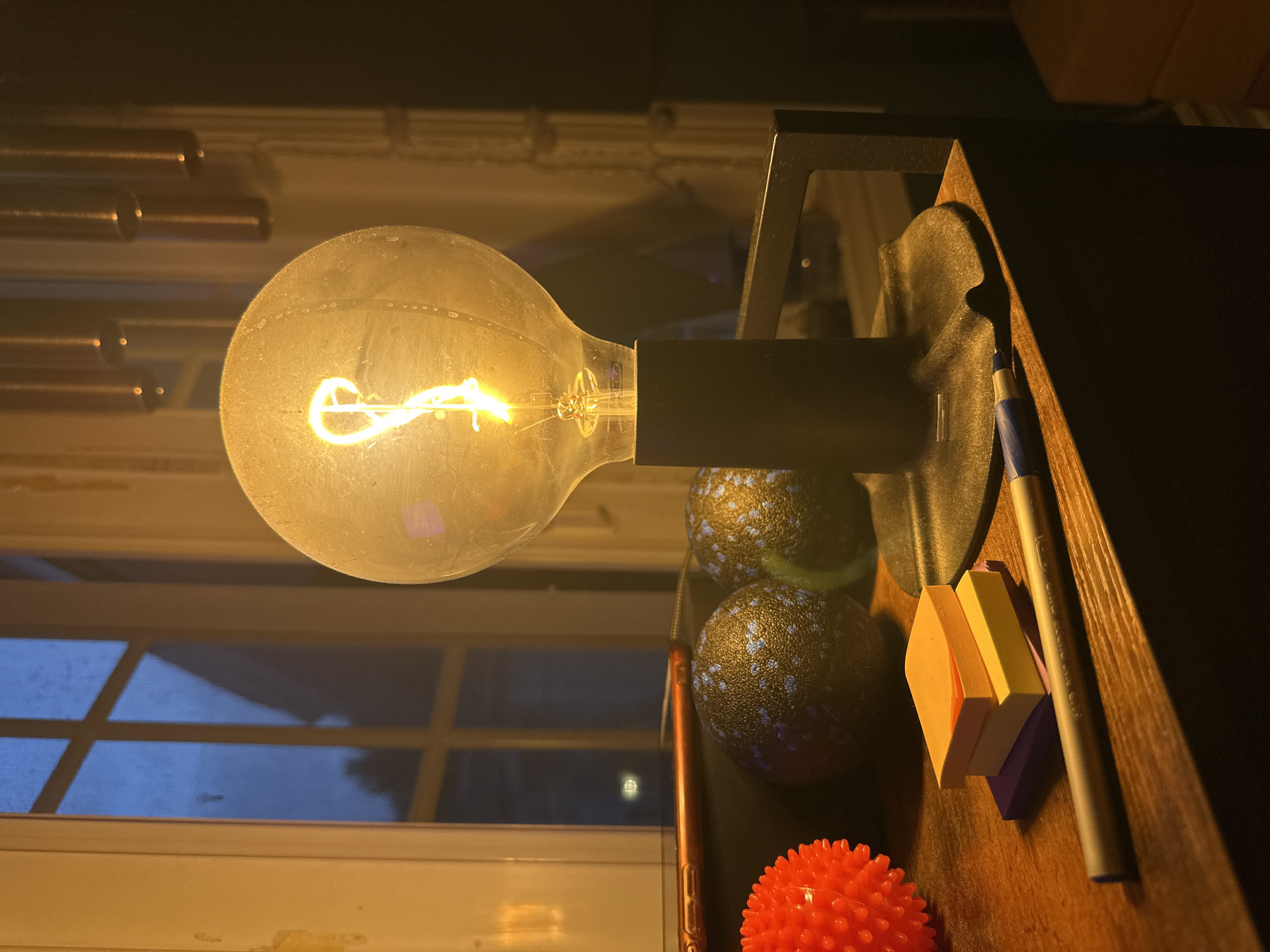 A close-up, low-angle shot captures a dimly lit room with a desk in the foreground. The focal point is a vintage-style incandescent lightbulb, its filament glowing warmly, casting a yellow light. To the left of the bulb are three stacks of colorful sticky notes, purple, yellow, and orange, along with a silver pen. Behind the sticky notes are two dark-colored, textured spheres and a vibrant red spiky ball. The background reveals a window with a grid pattern, reflecting a dark blue sky with hints of clouds. The room appears to be situated in an urban environment, possibly an apartment or house in McMasterville, Canada.