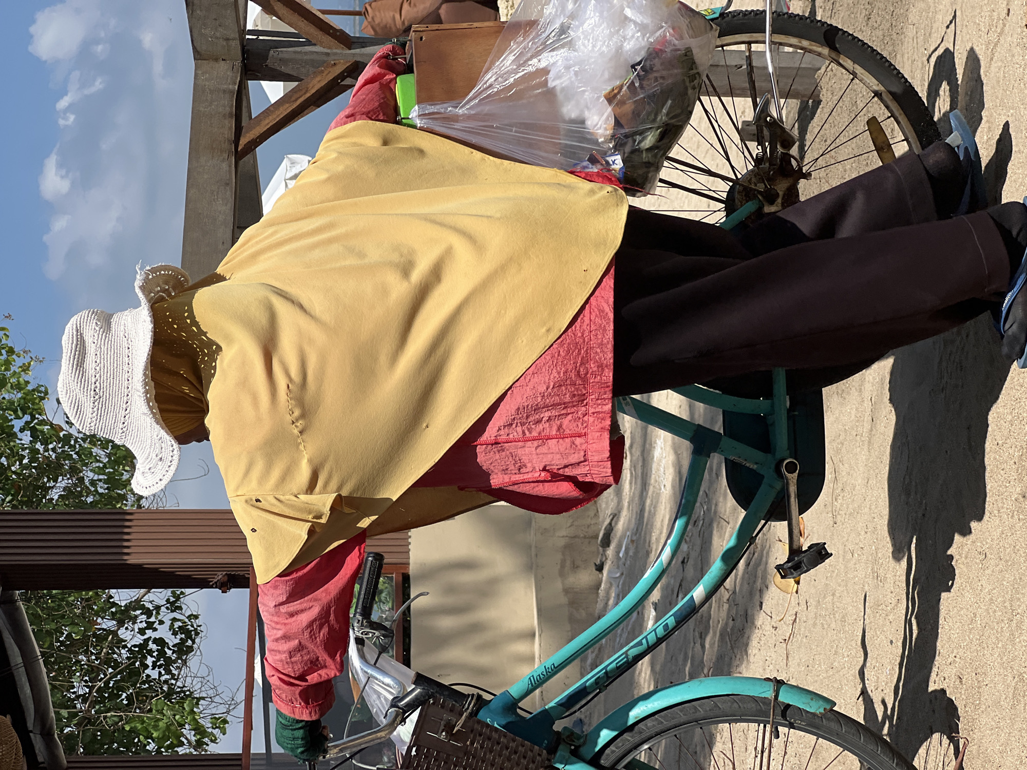 In Kuta, Indonesia, a person wearing a white hat, a yellow headscarf, a red long-sleeved shirt, and dark pants is seen from behind, standing next to a turquoise bicycle. The person is holding the bicycle's handlebars and has a large plastic bag filled with items attached to the back of the bicycle, as well as a wooden box. The scene appears to be outdoors, on a sandy surface, with a wooden structure and trees in the background. The sky is visible and appears to be partly cloudy.