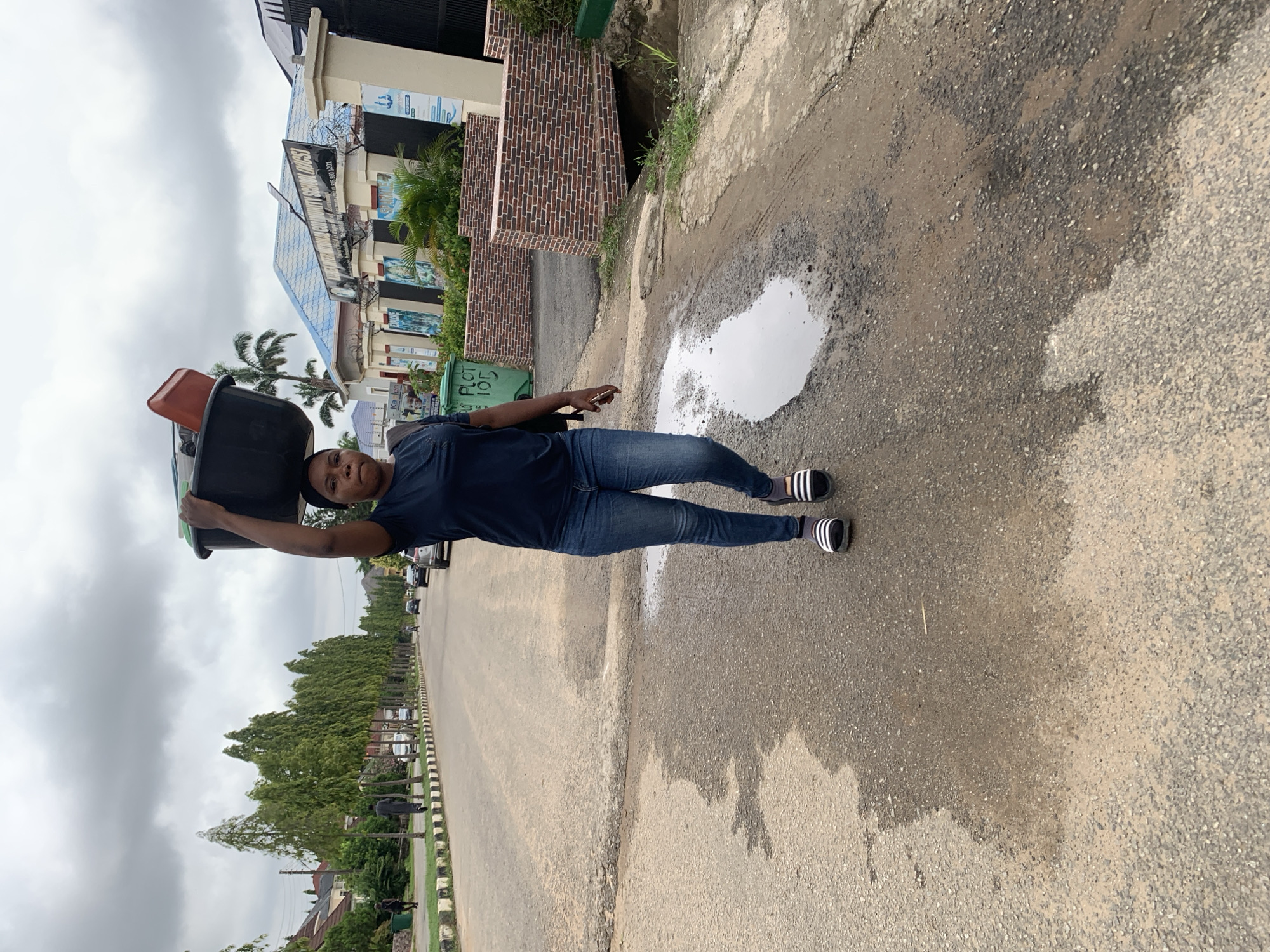A full, outdoor, medium shot captures a woman walking on a paved road in Abuja, Nigeria, carrying a black basin filled with various items, including a red container, on her head. The woman is wearing a dark blue t-shirt and blue jeans, with black and white striped slides on her feet. Her right hand is extended upward, supporting the basin. The road is wet with a large puddle in the foreground. To the left of the road, a row of tall, slender trees lines the street, with houses and buildings visible behind them. A sign on a building in the background reads "PLOT 105". The sky is overcast with gray clouds. The overall environment suggests an urban residential area in Abuja.