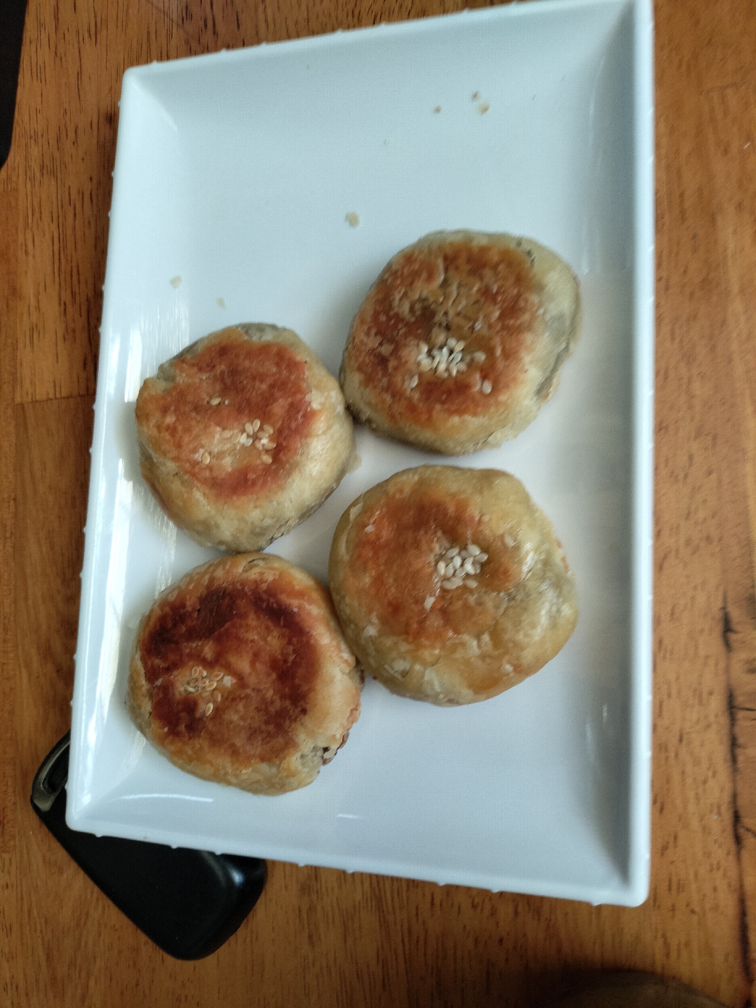 Four round, golden-brown pastries are arranged on a white rectangular plate, which sits on a wooden surface. Each pastry has a slightly uneven surface with areas of darker browning, indicating they have been baked or fried. A few white sesame seeds are scattered on the surface of each pastry, some forming small clusters. The pastries are positioned in a roughly square formation on the plate, with two in the top half and two in the bottom half. The wooden surface appears to be a table with a visible grain. The overall lighting suggests an indoor setting, possibly a restaurant or home kitchen. The image does not contain any information that would place it in the city of Langsa, Indonesia.