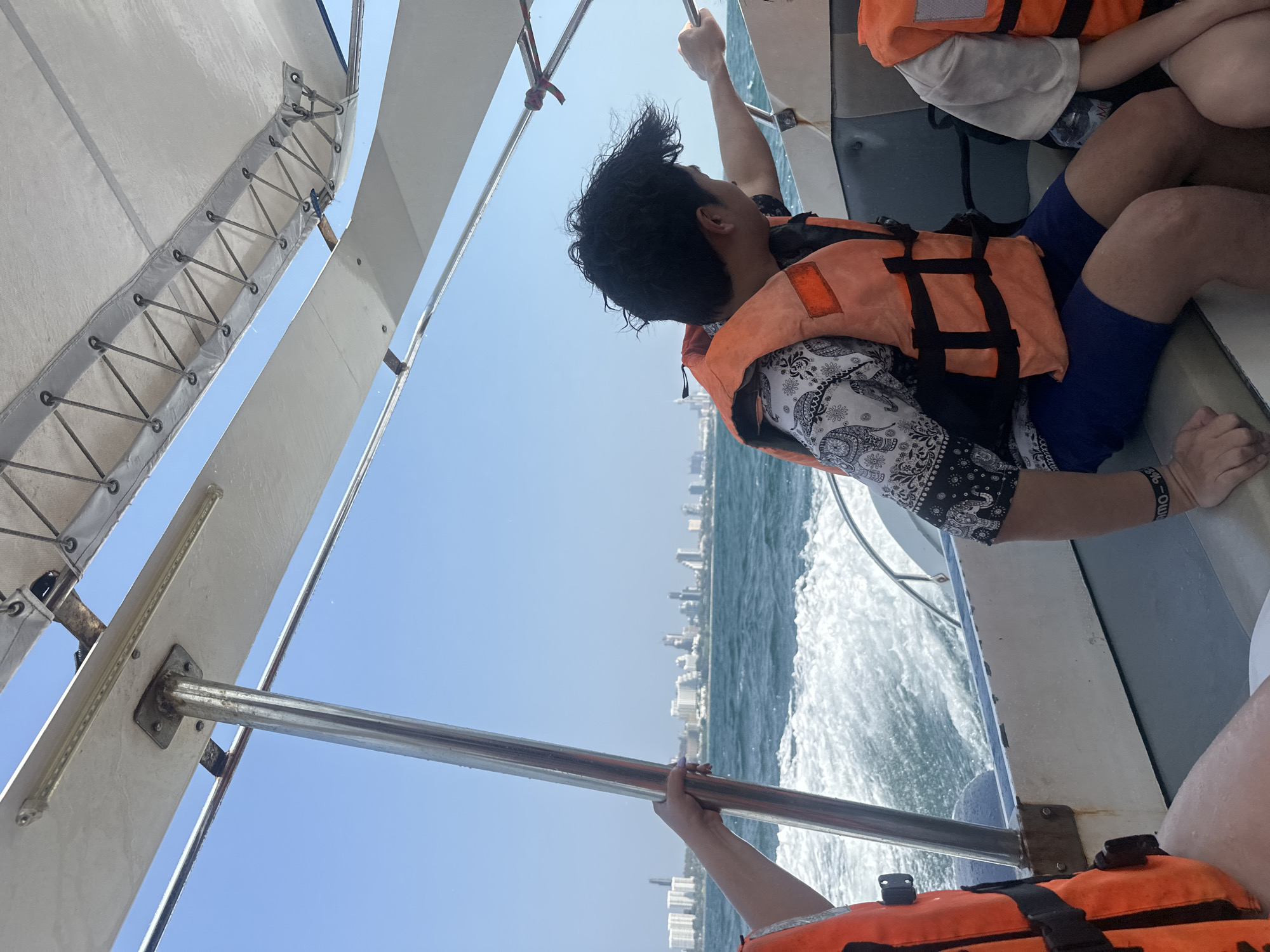 The image shows a group of people on a boat in Pattaya City, Thailand. One person with curly dark hair is wearing an orange life jacket and patterned shirt. The person is holding onto the boat's structure and looking towards the city skyline in the distance, which consists of many buildings. Another person is wearing an orange life vest and navy shorts. The boat is moving through the water, creating a wake visible behind it. The sky is blue and sunny. Parts of the boat's structure are visible in the foreground.
