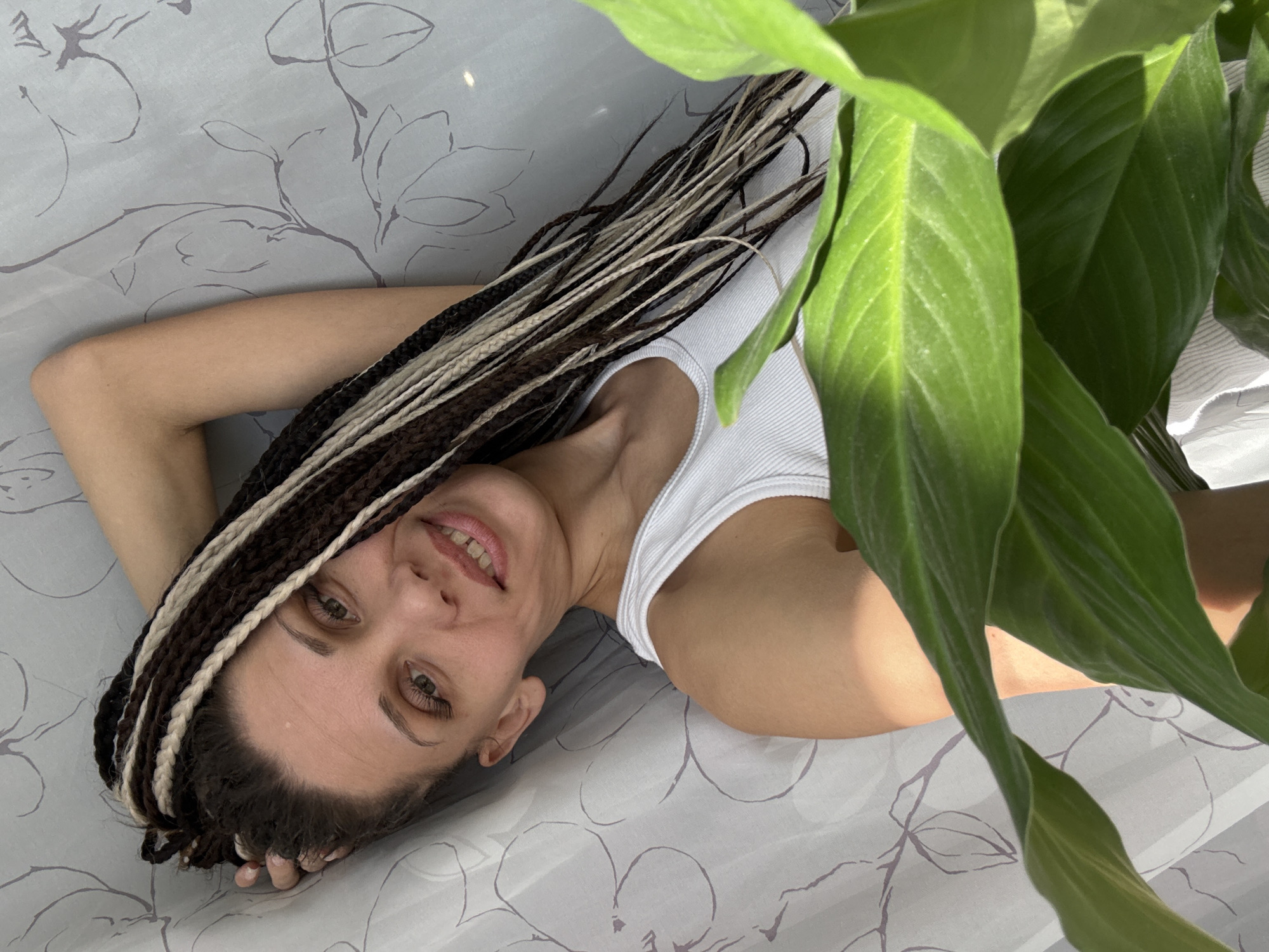 A young woman is depicted lying on her back, angled diagonally across the frame, against a light grey surface adorned with a subtle grey floral or leafy pattern. Her head is positioned towards the bottom right, and her body extends towards the upper left. She has braided hair, a mix of dark brown and blonde/light-colored strands. She is wearing a white ribbed tank top.
The woman is smiling gently, with her lips slightly parted, revealing her teeth, and she looks directly at the viewer with open eyes. Her expression is calm and inviting. Her left hand is visible near her head, resting on the patterned surface.
In the upper right portion of the image, a vibrant green plant with large, broad leaves partially frames the scene, obscuring part of her right shoulder and arm. The plant, possibly a peace lily, suggests an indoor setting. The bright, natural lighting indicates it is daytime. No text is visible. The scene suggests a relaxed moment in an indoor environment, potentially a home in Rusanovo, Russia.