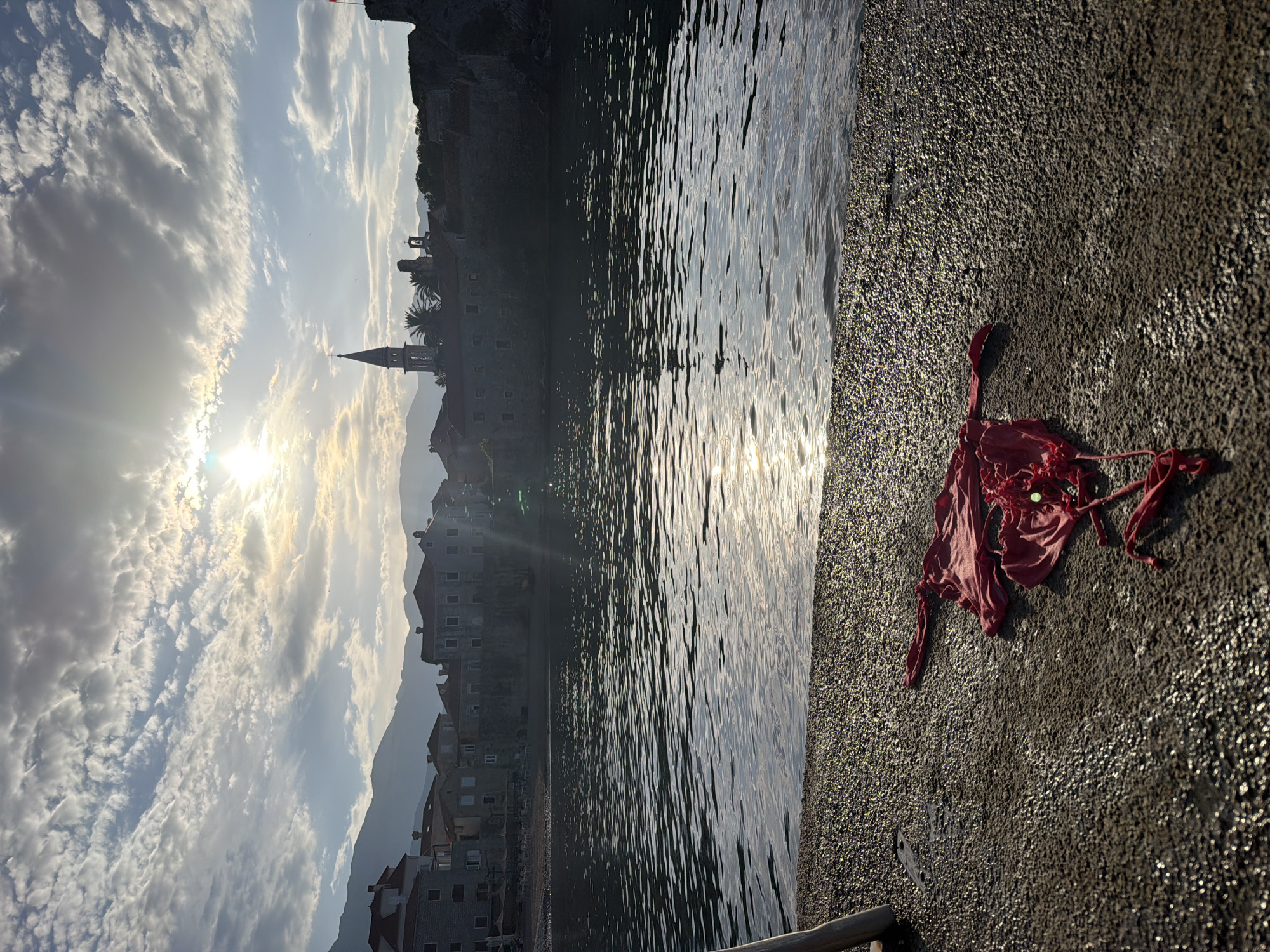 The image shows a seaside scene in Budva, Montenegro. The cityscape features old buildings with a prominent church spire under a sky with scattered clouds and a sunburst. The water reflects sunlight. On the right, a concrete surface, possibly a pier or seawall, is visible. A discarded piece of red cloth lies on the concrete. A partial railing is at the lower left.