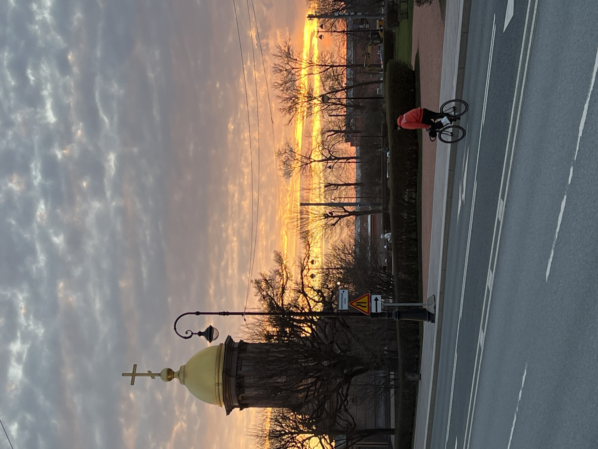 The image shows a sunset scene in Saint Petersburg, Russia, with a view of the Smolny Cathedral's golden dome in the foreground. The sky is filled with clouds reflecting the colors of the sunset, primarily orange and yellow hues. A bicyclist in a red jacket is riding along the street on the right side of the frame. The street has white lane markings. Bare trees line the boulevard, silhouetted against the bright sky. There is a street lamp and road signs visible along the side of the road.