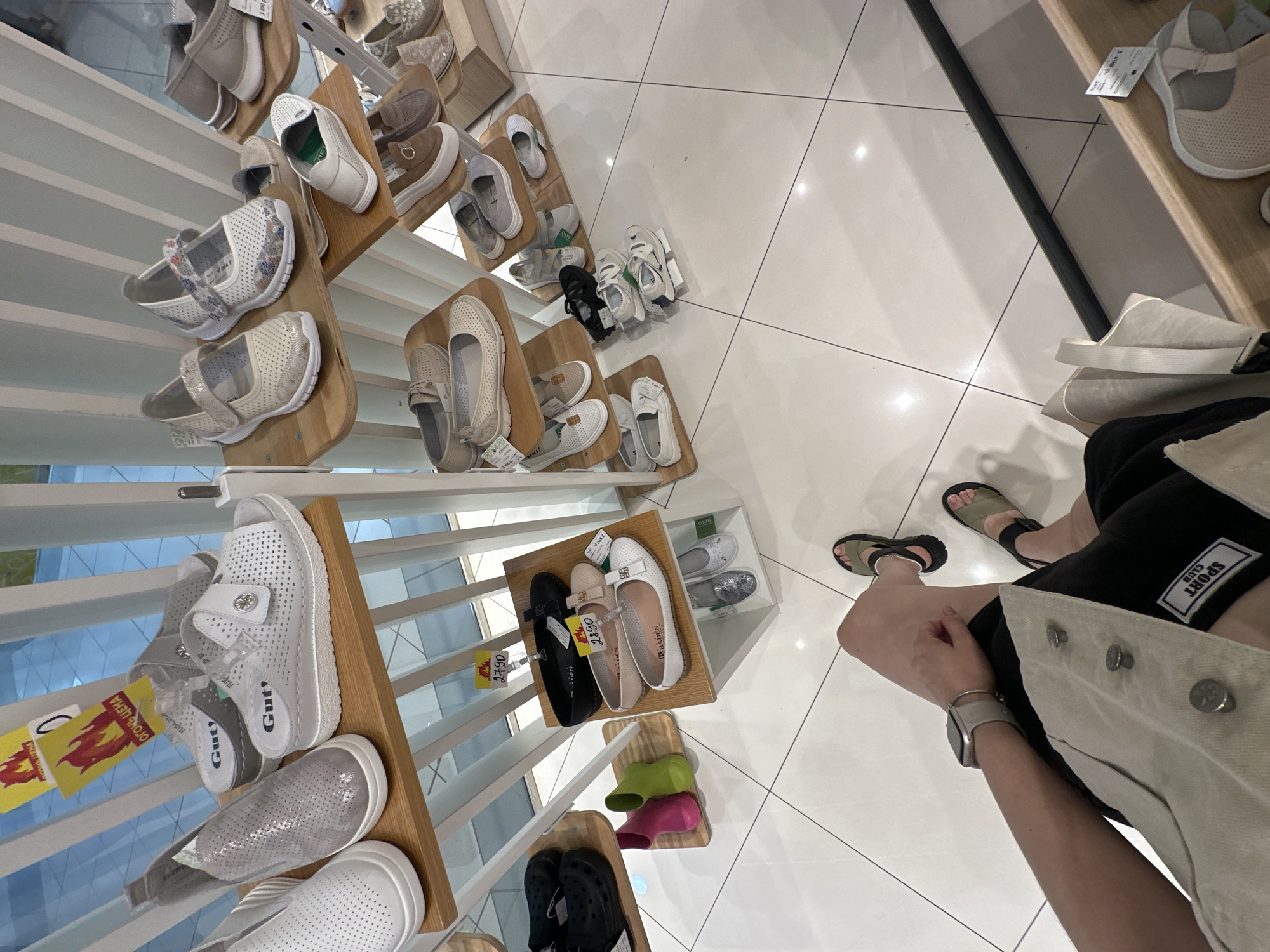 A high-angle shot depicts a shoe store in what is possibly Perm, Russia, evident from Cyrillic script on a tag attached to a pair of shoes. The camera is likely held by a person, whose hands, wrist with a smartwatch, lower leg, and feet clad in sandals are partially visible in the lower right. These body parts are positioned near shoe displays.
On the left and in the background are shelves containing multiple pairs of shoes, primarily women’s styles, arranged on wooden platforms. The shoes come in various colors, including white, gray, beige, black, pink, and green. They range from casual flats to sandals and closed-toe shoes, some featuring velcro straps.
The flooring is light-colored tile. The image conveys a sense of shopping and the variety of footwear available in a retail environment.