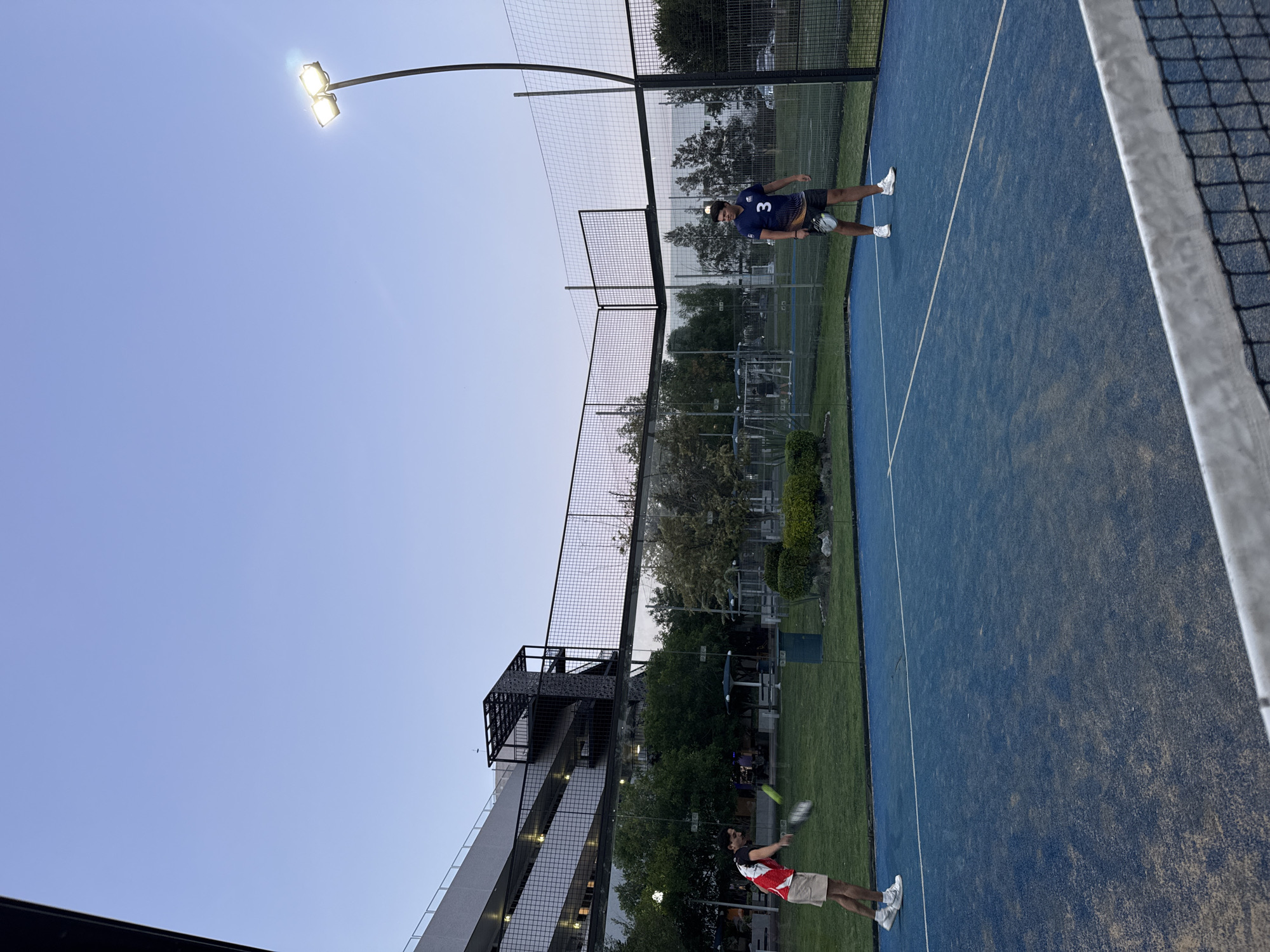 The image depicts two men playing padel tennis on a blue outdoor court in Puebla, Mexico, during dusk or early evening. The court features white boundary lines and is enclosed by tall mesh fences.
One player, located on the far side of the court, wears a blue short-sleeved shirt with the number "3" on the back, black shorts, and white athletic shoes, holding a padel racket. The second player, closer to the foreground, is dressed in a red and white short-sleeved shirt, beige shorts, and white athletic shoes. This player is captured mid-swing, seemingly hitting a blurred yellow padel ball. A white-taped padel net separates the two sides of the court.
Illuminating the scene are two bright floodlights mounted on a tall pole, indicating the fading natural light. In the background, beyond the court's fencing, a large, multi-story building, possibly a parking garage or sports complex facility, with visible metal structures, rises against a clear, pale blue sky. Lush green grass and trees surround the court, suggesting an urban or suburban sporting environment. The overall atmosphere is focused on the ongoing game.