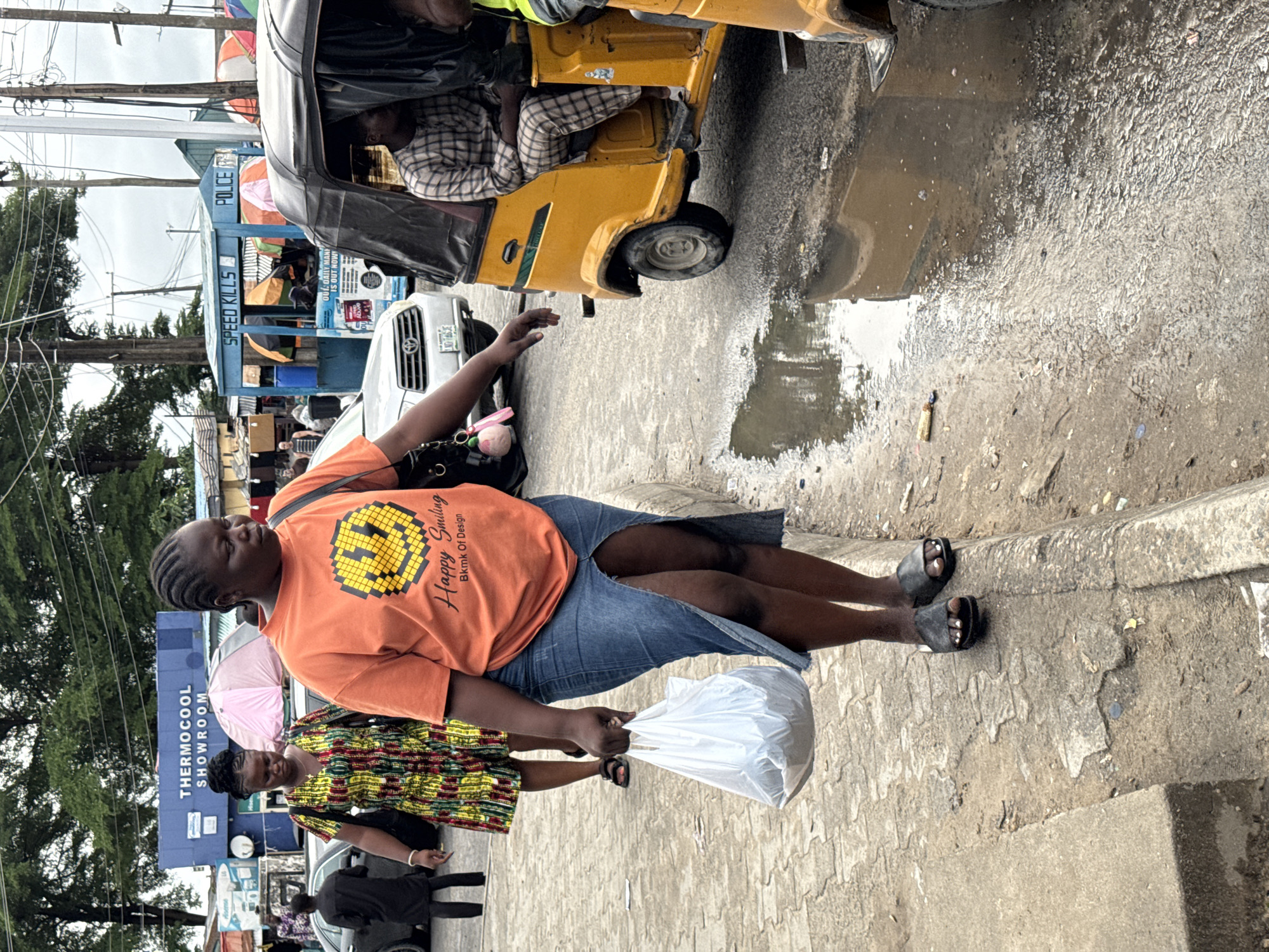 The image shows a street scene in Ado, Nigeria. A woman wearing an orange "Happy Smiling" t-shirt and a denim skirt stands on the sidewalk, holding a white plastic bag. Another woman in a colorful patterned dress stands beside her. In the background, there's a blue "THERMOCOOL SHOWROOM" sign and a blue police booth that says "SPEED KILLS" and "POLICE" on it. A yellow auto rickshaw (tuk-tuk) is partially visible in the upper right corner of the frame, parked near a large puddle. A white car is parked behind the police booth. Several people are walking around in the background, and power lines are visible overhead.