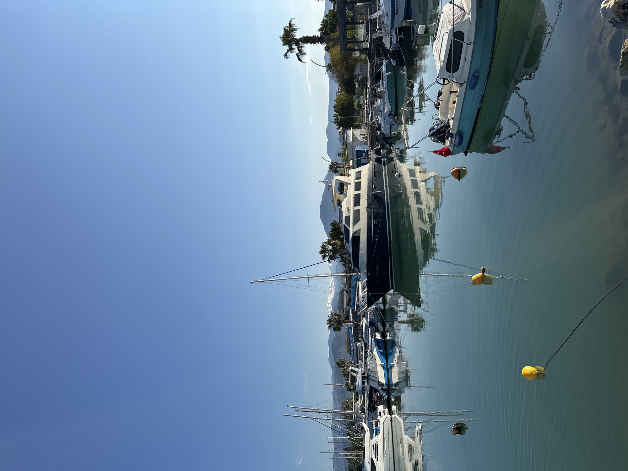 The image depicts a marina in Fethiye, Turkey. Several boats of varying sizes are docked along a pier, their reflections visible in the calm water. Mountains are seen in the background, with palm trees interspersed along the shoreline. The sky is clear and blue. The boats are secured with mooring lines and some have buoys attached.