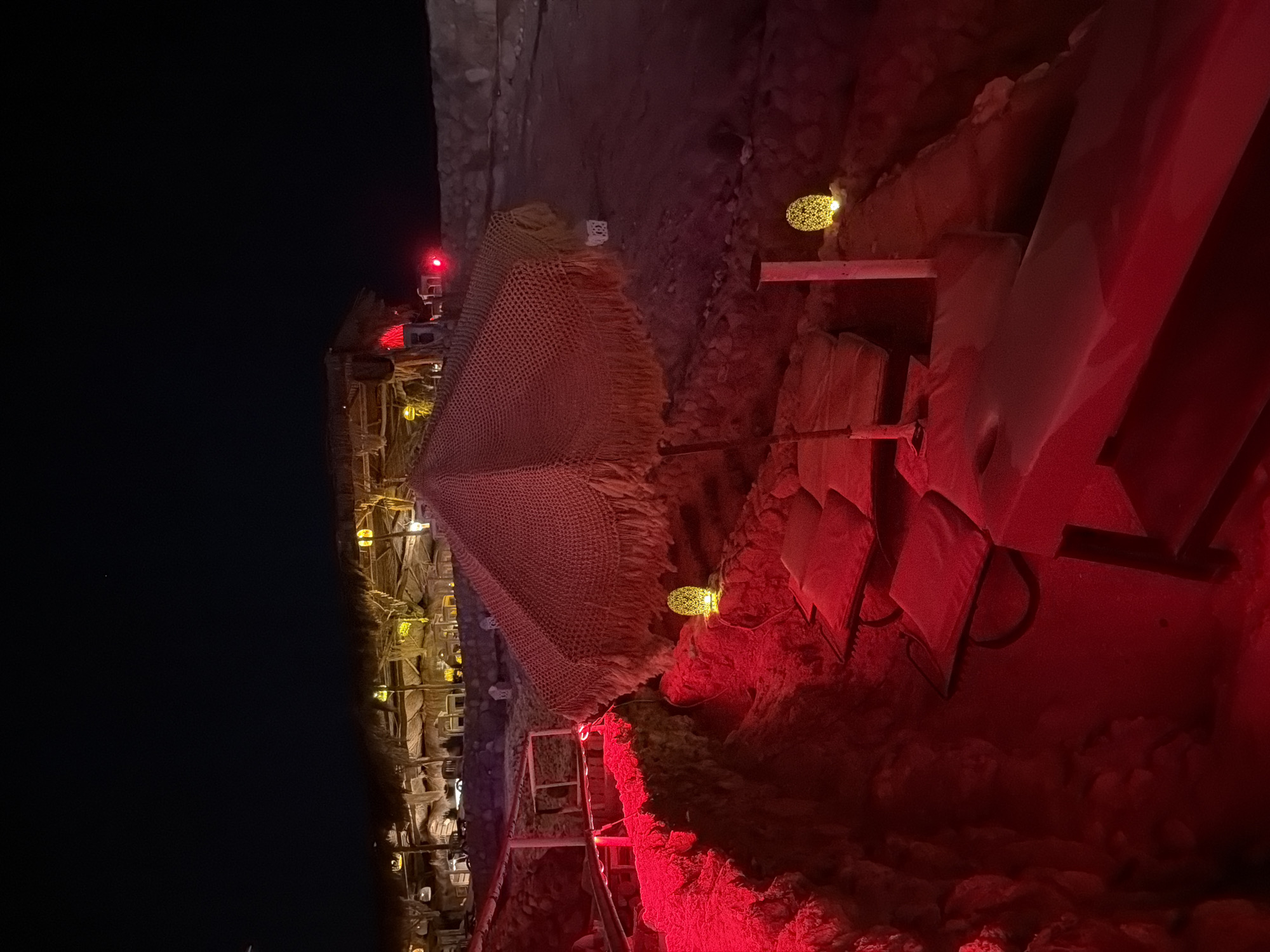 A nighttime outdoor scene in Sharm El Sheikh, Egypt, depicts a beachfront lounge area illuminated by red lighting and decorative yellow lanterns. The scene features several lounge chairs and cushions arranged on tiered levels, some covered with a red fabric. A large, fringed umbrella with a thatched roof stands prominently in the foreground, casting shadows. Beyond the lounge area, a building with a thatched roof and exposed beams is visible, with interior lights creating a warm glow. Small red lights are also present, possibly indicating signage or safety markers. The ground appears to be sandy or rocky, typical of a beach environment. The overall ambiance is relaxed and inviting, suggesting a resort or restaurant setting.