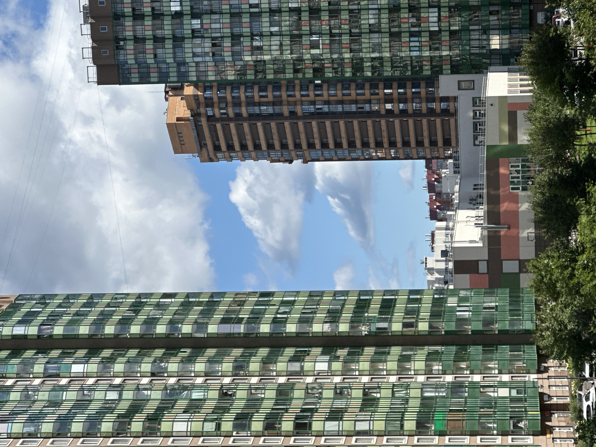 The image depicts a section of a modern urban residential area, likely in Russia, given the architectural style. Dominating the frame are two tall, multi-story apartment buildings. The building on the left is characterized by its facade covered in green vertical louvers and numerous glass-enclosed balconies, giving it a distinctive striped appearance. To its right, a second, taller building features a facade of alternating green and reddish-pink square glass panes, reflecting the sky and clouds. Between these two prominent structures, a narrower, taller building constructed of reddish-brown brick with numerous windows is visible. In the foreground, a lower-rise building with a white and colored facade, possibly a community center or commercial space, is partially obscured by a dense canopy of green trees. A few cars are parked on a street in the lower left corner, and overhead, power lines crisscross against a sky filled with scattered white clouds. The overall scene conveys a sense of contemporary urban development with a focus on residential complexes.