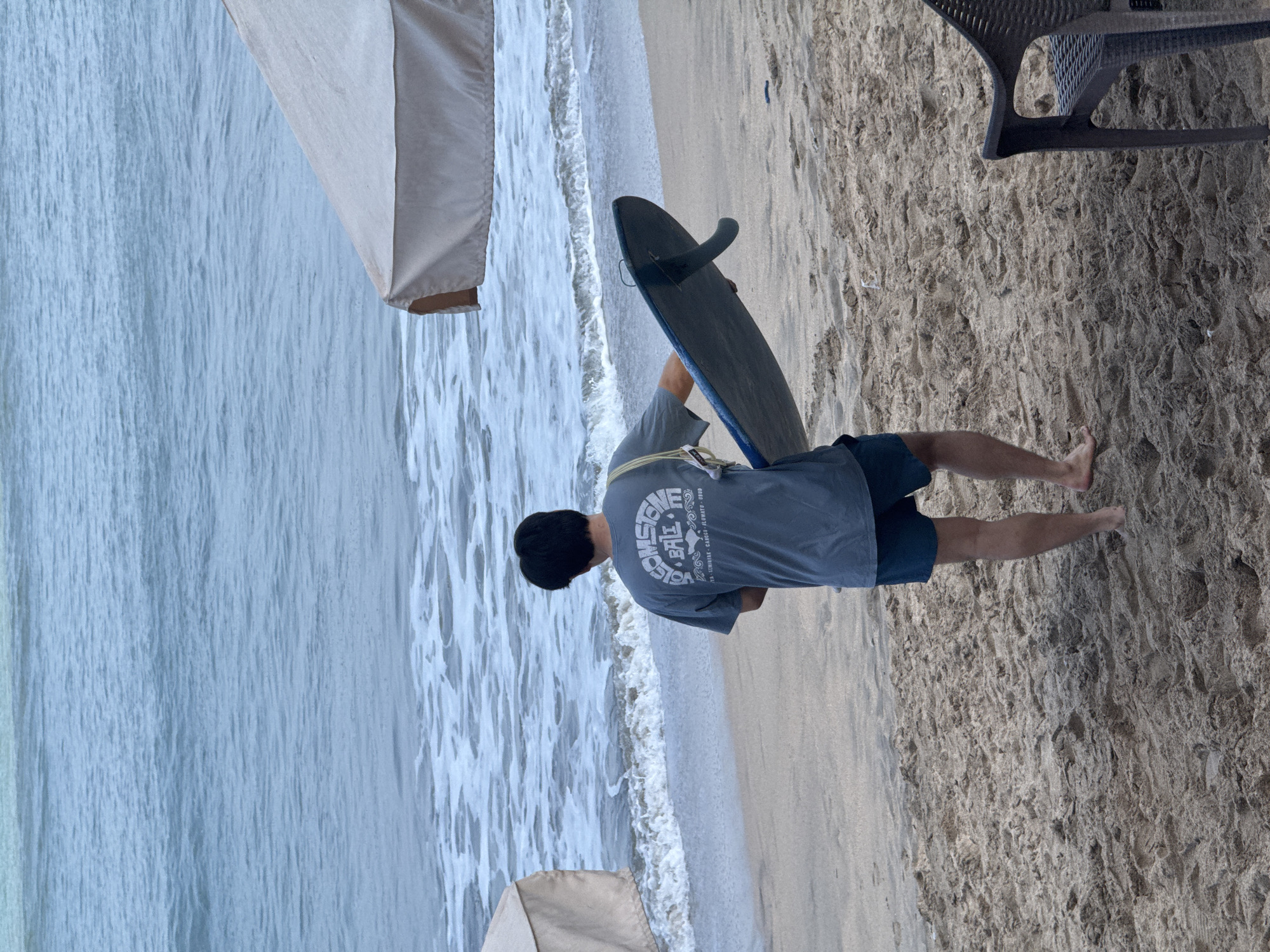 A person is standing on a sandy beach in Kuta, Indonesia, holding a blue surfboard. They are facing away from the camera, towards the ocean, with waves breaking on the shore. The person is wearing a grey t-shirt and blue shorts. In the background, there are two large, light-colored umbrellas providing shade. To the right of the frame, a dark plastic chair is partially visible. The sand is textured with footprints and debris. The water is a muted blue-grey, with white foam from the waves.