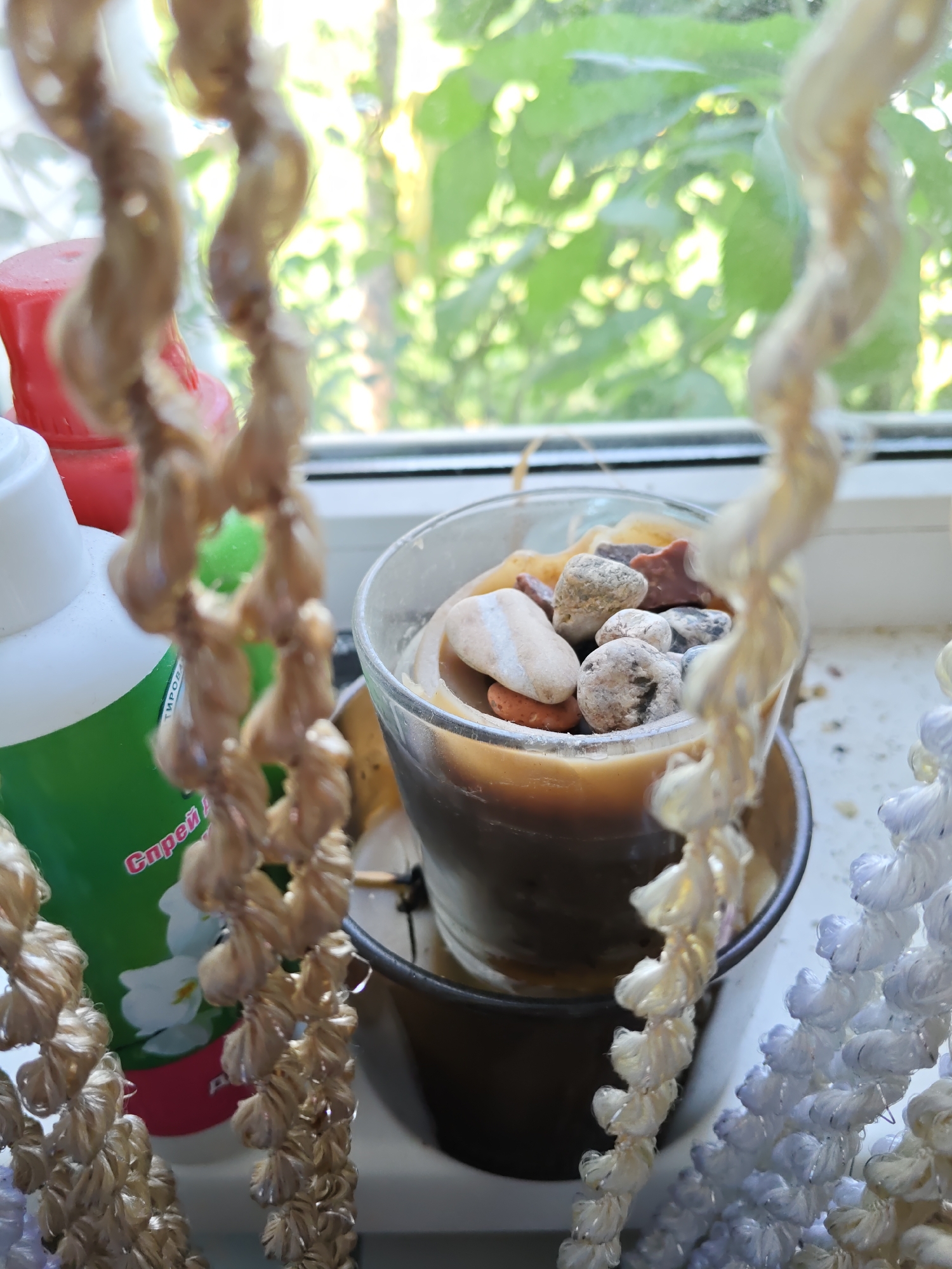 This is a close-up, slightly out-of-focus shot of a candle with small stones embedded in its wax. The candle is in a glass holder, which is sitting on a windowsill. To the left of the candle is a white bottle with a green label that has some text in Cyrillic. Behind the bottle and candle is a blurred background of green leaves, suggesting an outdoor environment visible through the window. In the foreground, two decorative strands made of braided material hang down, obscuring parts of the scene. One strand is a light brown color, and the other is white and light blue. The lighting suggests it is daytime, likely with natural light coming from the window. The location of Rostov-on-Don, Russia, is provided as context but is not visually discernible from the image itself.