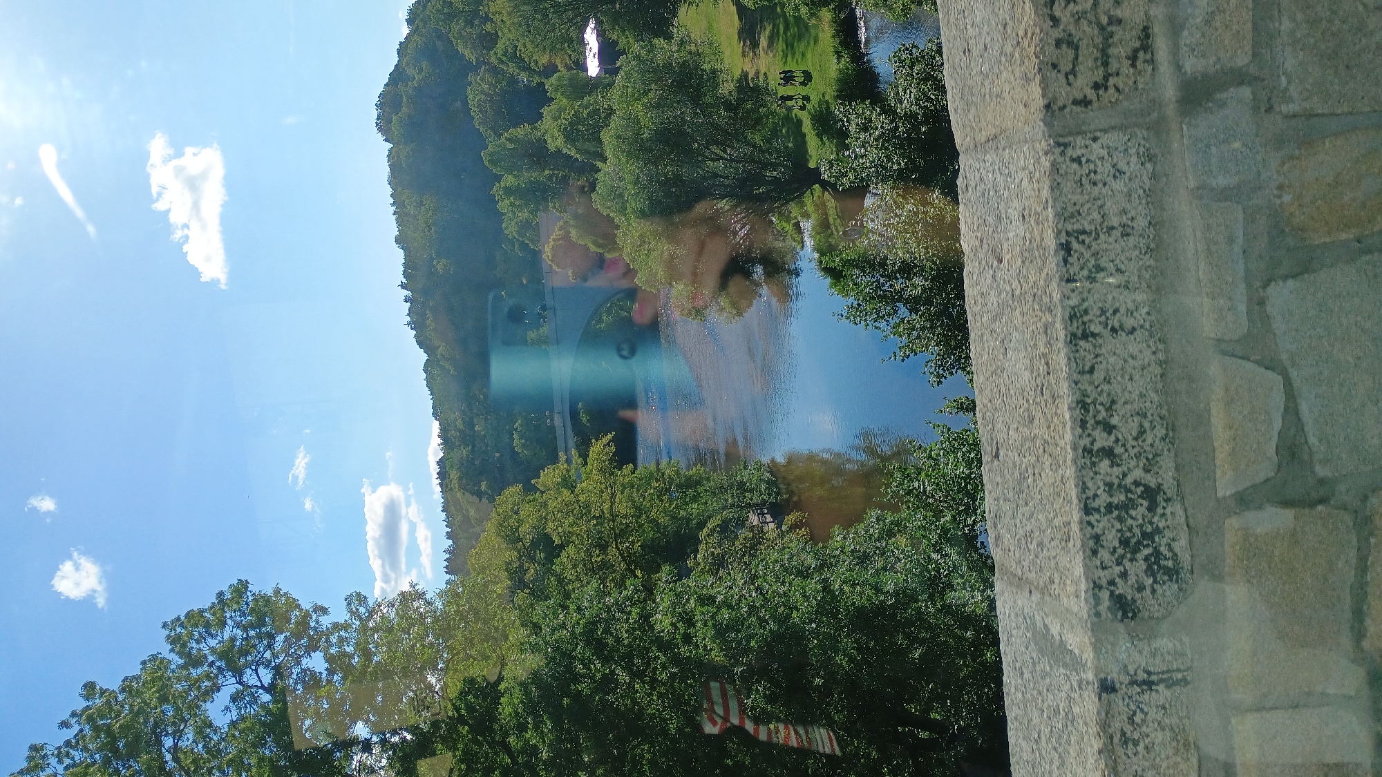 The image shows a wide river with a stone bridge spanning across it in the distance. The river is bordered by lush green trees on both sides, with a gentle hill covered in foliage forming the background. The sky is a vibrant blue with a few white clouds and the sun is visible, casting a bright glare on the water and sky. In the foreground, a stone wall made of large, irregular blocks is visible, suggesting it is part of a bridge or elevated structure. On the right bank of the river, a grassy area is visible with a small group of people gathered. The overall scene depicts a tranquil natural landscape with architectural elements.