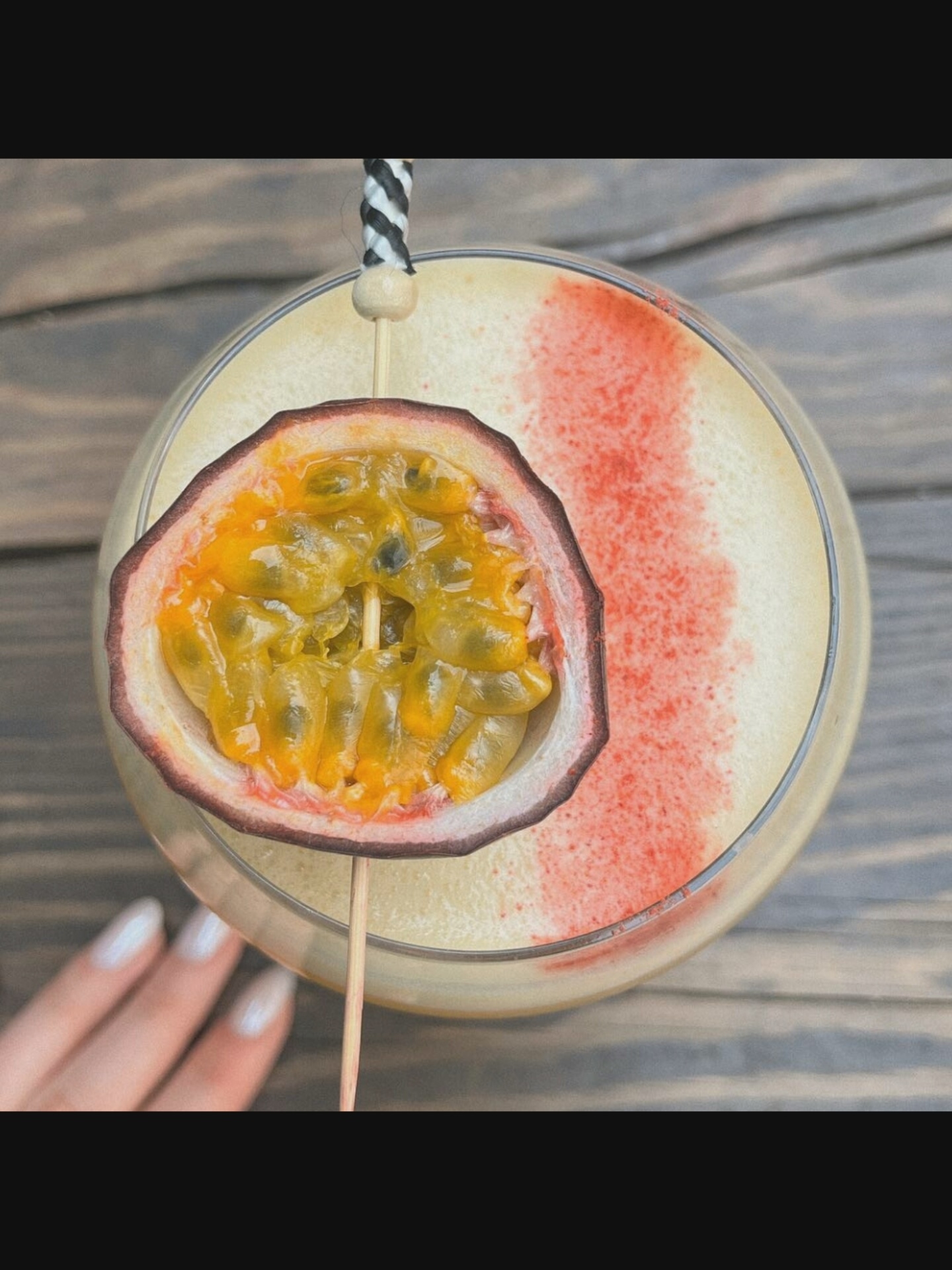 A close-up, overhead shot shows a cocktail in a clear glass, garnished with a slice of passion fruit and a black and white striped straw. The drink has a frothy, pale yellow surface with a dusting of red powder in a curved pattern. The glass sits on a rustic wooden table. A woman's hand with manicured nails is visible in the lower left corner, reaching towards the glass. The background is blurred, suggesting an outdoor setting. The scene is located in Bielawska Wieś, Poland.