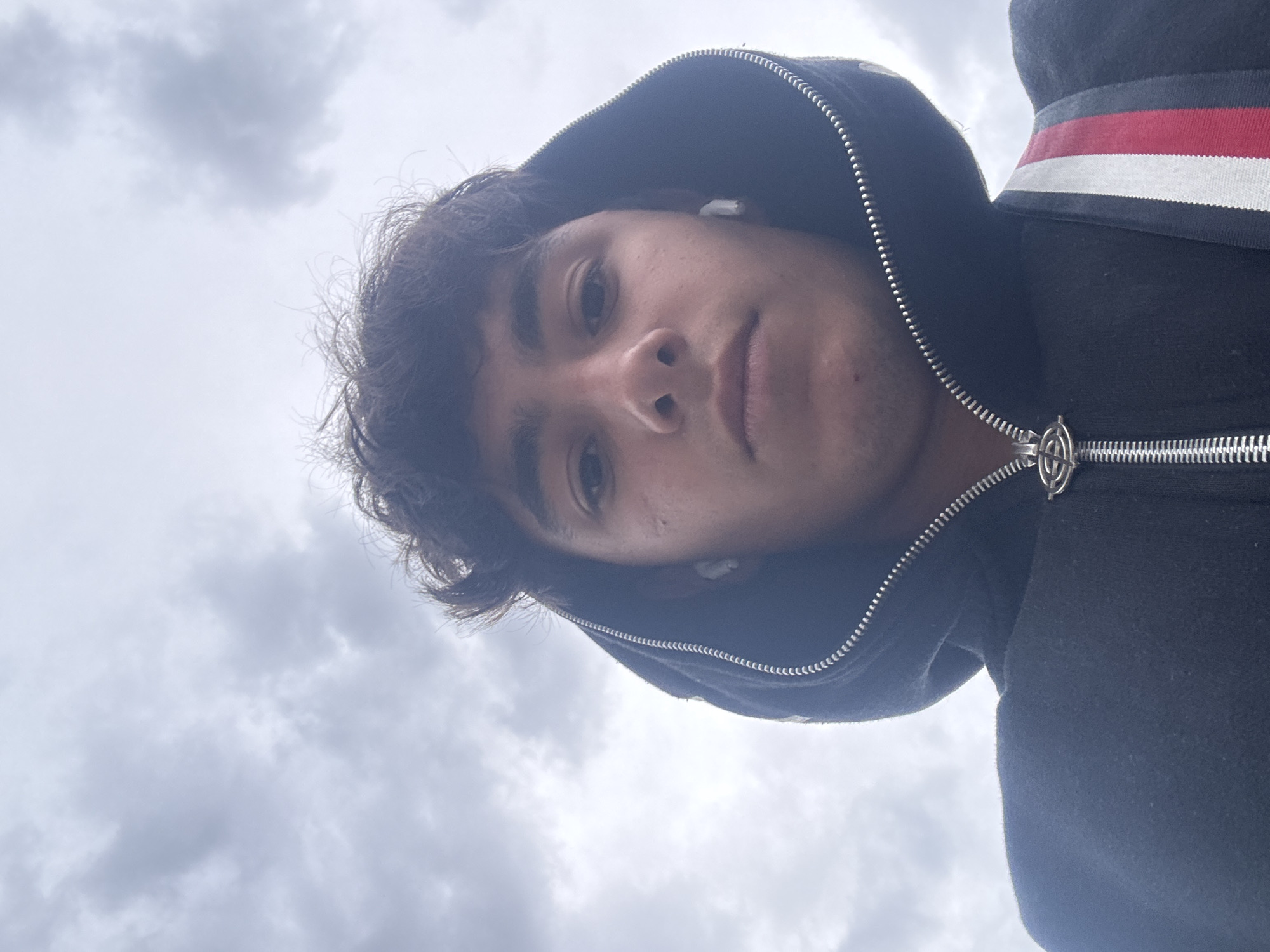 A young man is pictured from a low angle, possibly a selfie. He is wearing a black hooded sweatshirt zipped partially open, with white earbuds in both ears. The hood is up, framing his face. A portion of a backpack strap with red, white, and navy colors is visible over his left shoulder. The background is a cloudy sky. The location in Yerevan, Armenia is not apparent from the image.
