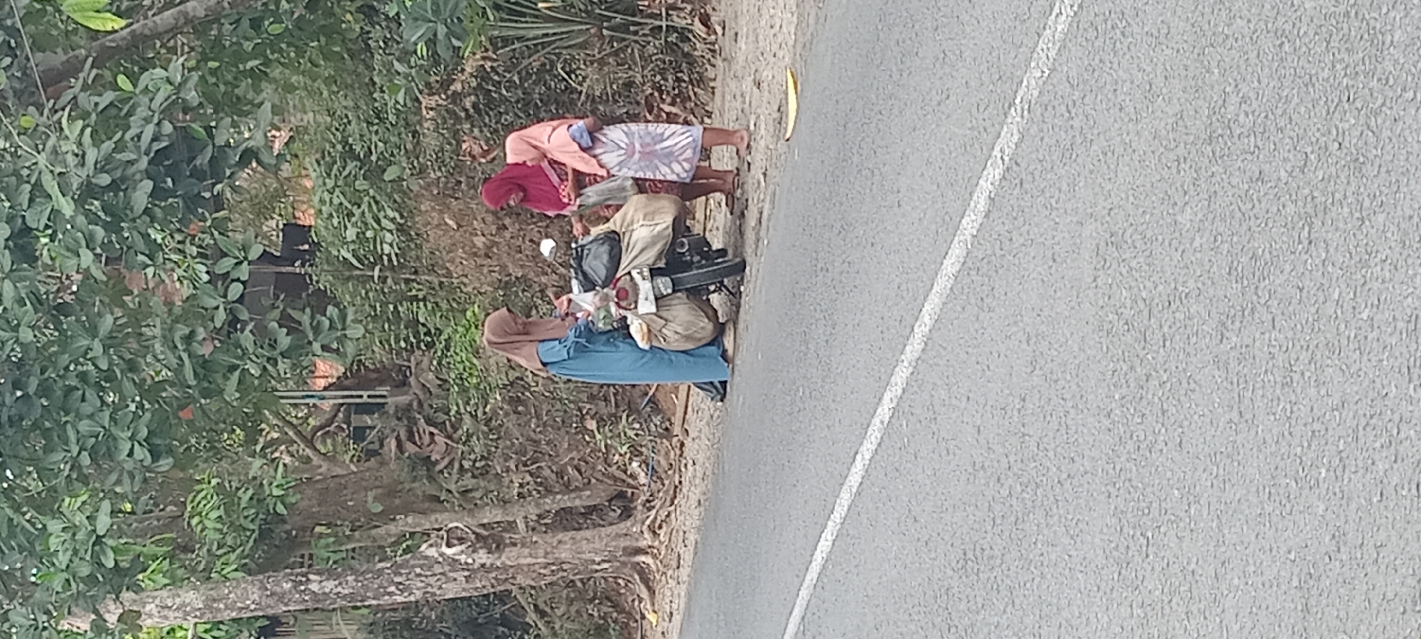 Three individuals, appearing to be women dressed in traditional Islamic headscarves and modest clothing, are gathered around a motorcycle on the side of a paved road. The motorcycle is parked and loaded with several large bags, suggesting it is used for transport or carrying goods. One woman, wearing a blue dress and a brown headscarf, is on the left, interacting with items on the motorcycle. Another woman, in a red headscarf, is standing in the middle, also seemingly engaged with the motorcycle's contents. A third woman, wearing a pinkish headscarf and a patterned skirt, stands to the right, observing or interacting with the others.
The scene is set outdoors, with a roadside environment characterized by a paved road with a white line marking, a sandy or dirt shoulder, and dense vegetation including trees and bushes. The background shows a sloping embankment covered in greenery and some visible man-made structures or debris. The overall impression is a rural or semi-rural setting, likely in Cimaragas, Indonesia, given the presence of vegetation and the style of dress. The lighting suggests it is daytime, with natural light illuminating the scene.