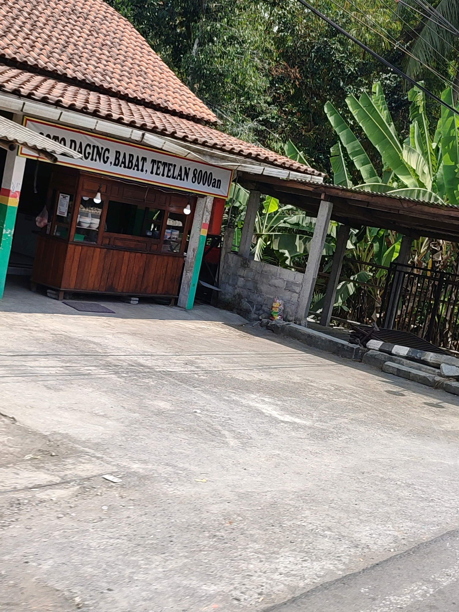 A food stall named "Soto Daging Babat Tetelan 8000an" is situated in Sawangargo, Indonesia. The stall features a wooden counter with visible bowls of food. Above the counter, a banner displays the stall's name in red and black text against a white background, bordered by yellow. The stall is covered by a tiled roof. To the right of the stall, a covered outdoor seating area is supported by concrete pillars. Beyond this area, a black metal fence separates the property from lush vegetation, including banana trees, and overhead power lines are visible. The ground in front of the stall and extending to the foreground is paved with concrete. A small, colorful pile of items, possibly trash bags, is located near the base of the concrete pillars.
