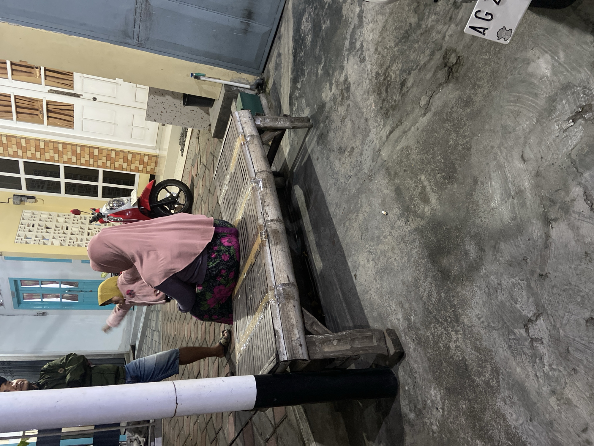 The image captures an outdoor scene in Tulungagung, Indonesia, at what appears to be dusk or early evening. In the foreground, a rustic bamboo bench is positioned on a concrete surface that shows signs of wear and tear. The bench is partially covered with a patterned cloth, suggesting it might be used for seating.
To the left of the bench, a white cylindrical pole, possibly a lamp post, stands prominently. Behind the bench, a woman wearing a pink hijab and a floral skirt is seated. Her body is angled away from the camera, and she appears to be interacting with a child. The child, visible between the woman and another adult, is wearing a yellow head covering and a pink jacket, with their arm extended. To the far left, an adult male, dressed in a dark jacket and denim shorts, stands with his arms crossed, facing the woman and child.
In the background, a red and white scooter is parked near a building. The building features a yellow facade with white-trimmed windows and a white double door. A blue-colored door is also visible further down the building. The ground is paved with interlocking bricks in some areas and concrete in others. The overall impression is of a casual gathering or a moment of rest outside a residential or commercial space in Tulungagung.