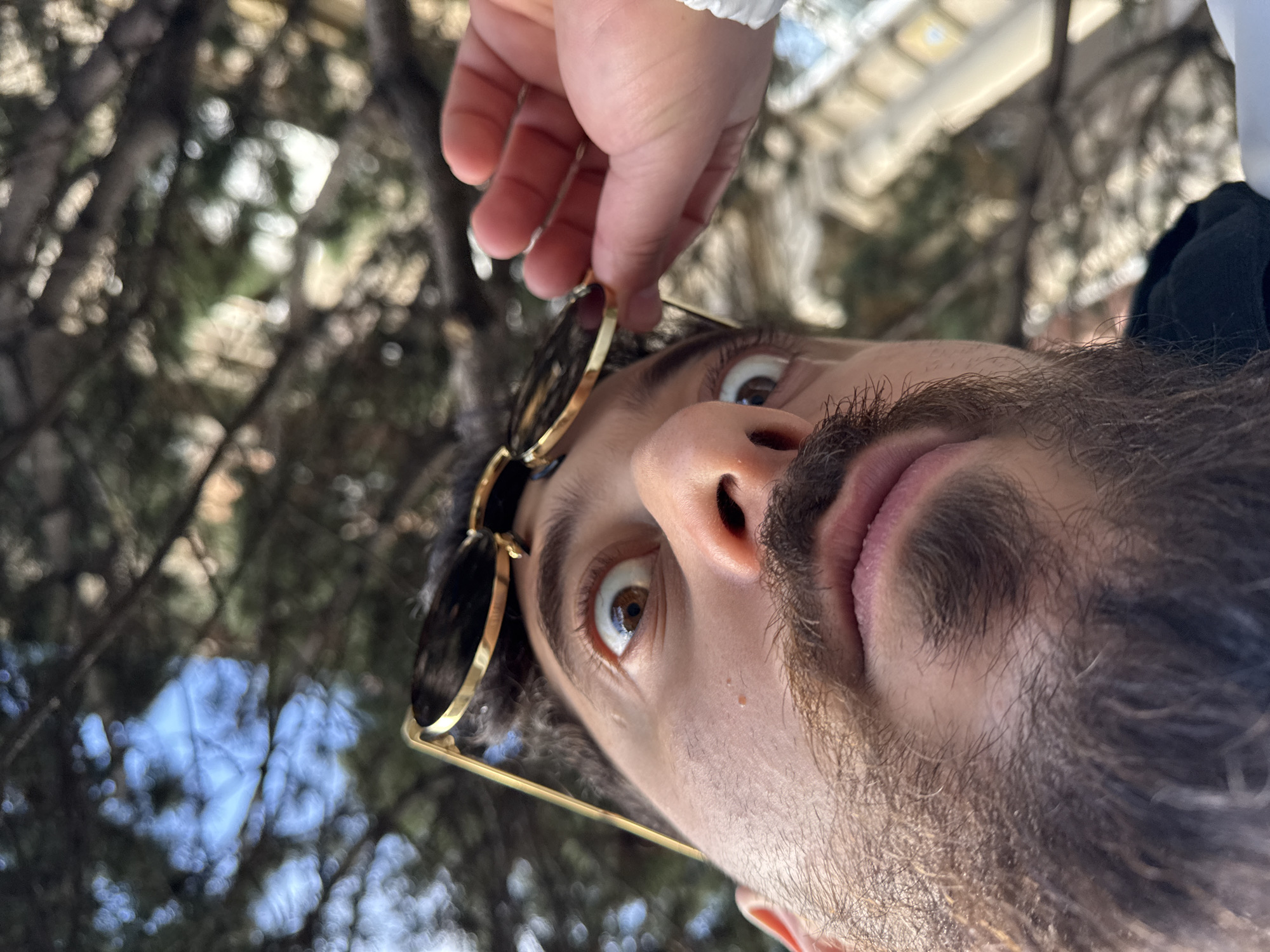 The image presents an adult male captured from a low-angle perspective, looking directly upwards. His face, featuring a dark beard and mustache, occupies the foreground. His brown eyes are wide, with visible whites and dilated pupils, fixed on an upward point. His mouth is slightly ajar, exposing a portion of his tongue. Round sunglasses with gold-colored metal frames rest on his forehead, partially held by his right hand (viewer's left), which is visible emerging from a white sleeve or cuff. The background is out of focus, showing a dense canopy of dark green tree foliage and intermittent patches of blue sky. Indistinct, blurred architectural elements are faintly discernible in the upper right quadrant. This scene is located in Strasbourg, France.
