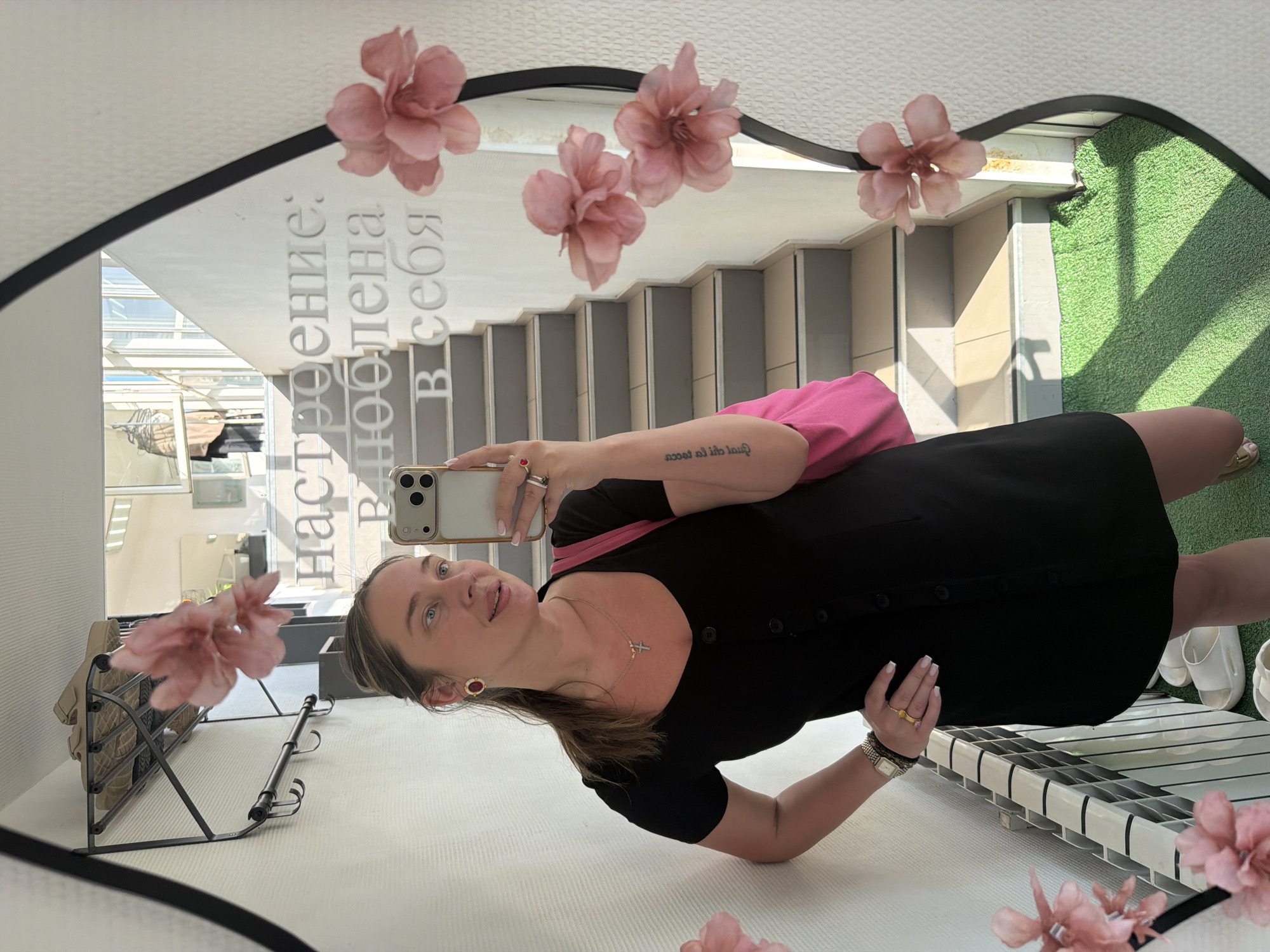 A woman stands in front of an ornate mirror in what appears to be a shop or commercial space in Chaikovsky, Russia. She is taking a selfie with her iPhone. The woman is wearing a black dress with a pink bag slung over her shoulder, a necklace with a cross, earrings, and a watch. The mirror has pink flower decorations and Russian text. In the reflection, there are stairs, green turf or carpeting, and various interior elements.