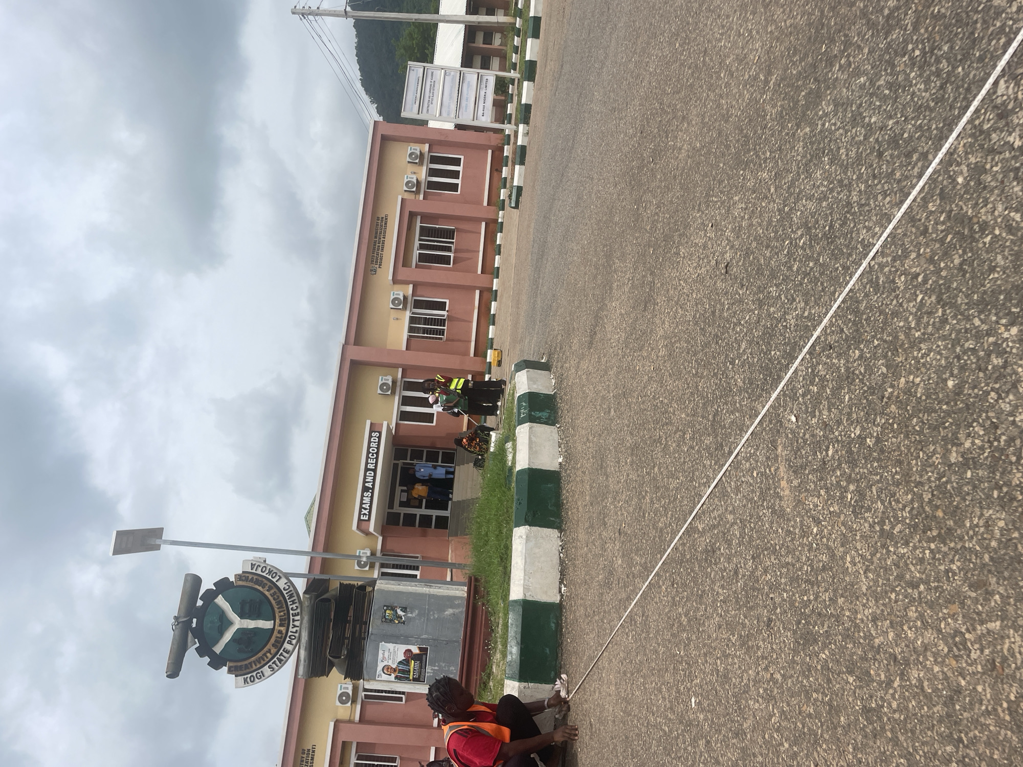 The image depicts the exterior of a building at the Kogi State Polytechnic, Lokoja, Nigeria. The building has a salmon-colored facade with a sign above the entrance reading "EXAMS. AND RECORDS". Several individuals are visible, including a person in an orange vest and black pants kneeling on the asphalt in the foreground, using a measuring tape. Other people are near the building entrance and on the sidewalk. A large emblem of the polytechnic is prominently displayed on a monument near the building, and a street light is positioned nearby. The environment includes paved ground, a grassy area, and trees in the background, with a cloudy sky overhead. The image appears to have been taken outdoors during the daytime.