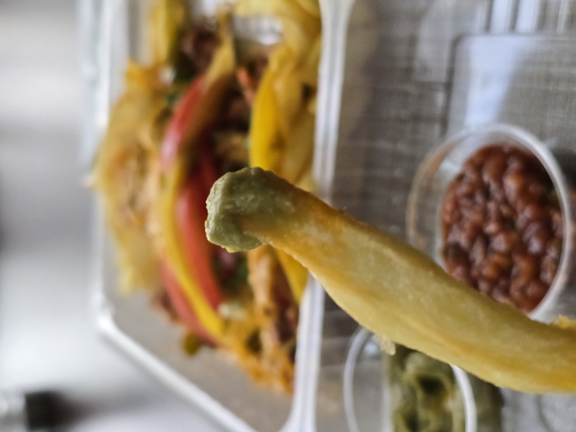A close-up shot shows a french fry dipped in a green sauce, held in the foreground. To the right are two small, clear plastic containers, one containing a red sauce and the other more of the green sauce. Behind the french fry and dipping sauces, inside a clear plastic container, is what appears to be the remainder of a dish, containing what looks like peppers and other food items. The setting is in Unknown, Unknown.